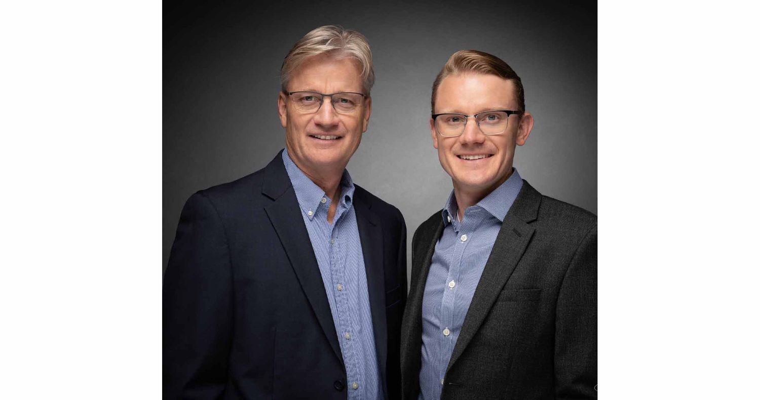 Two smiling men in glasses, dark blazers and blue collared shirts pose together against a dark gray studio background.