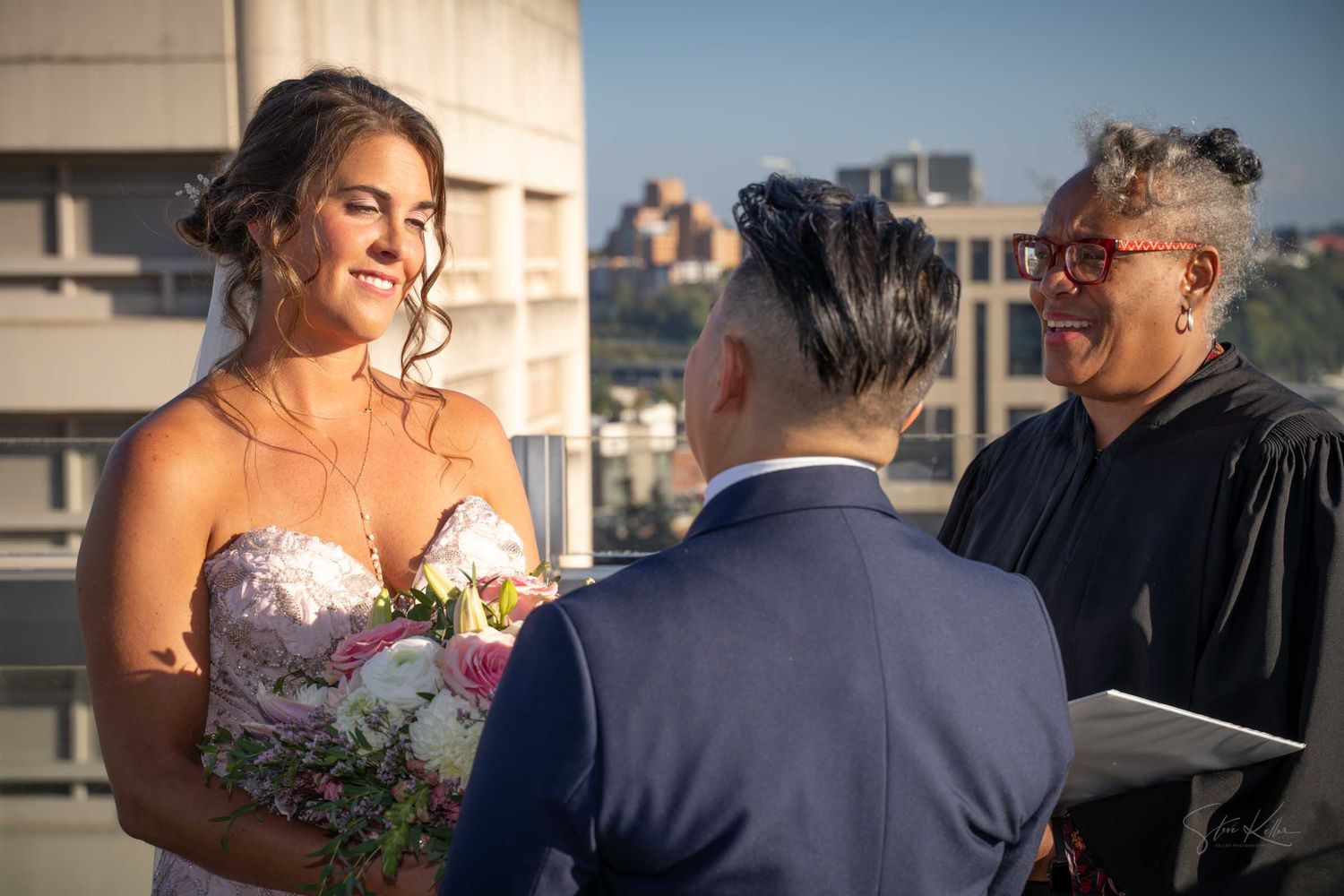 Smiling bride exchanges vows with groom as officiant reads from book during outdoor rooftop ceremony.
