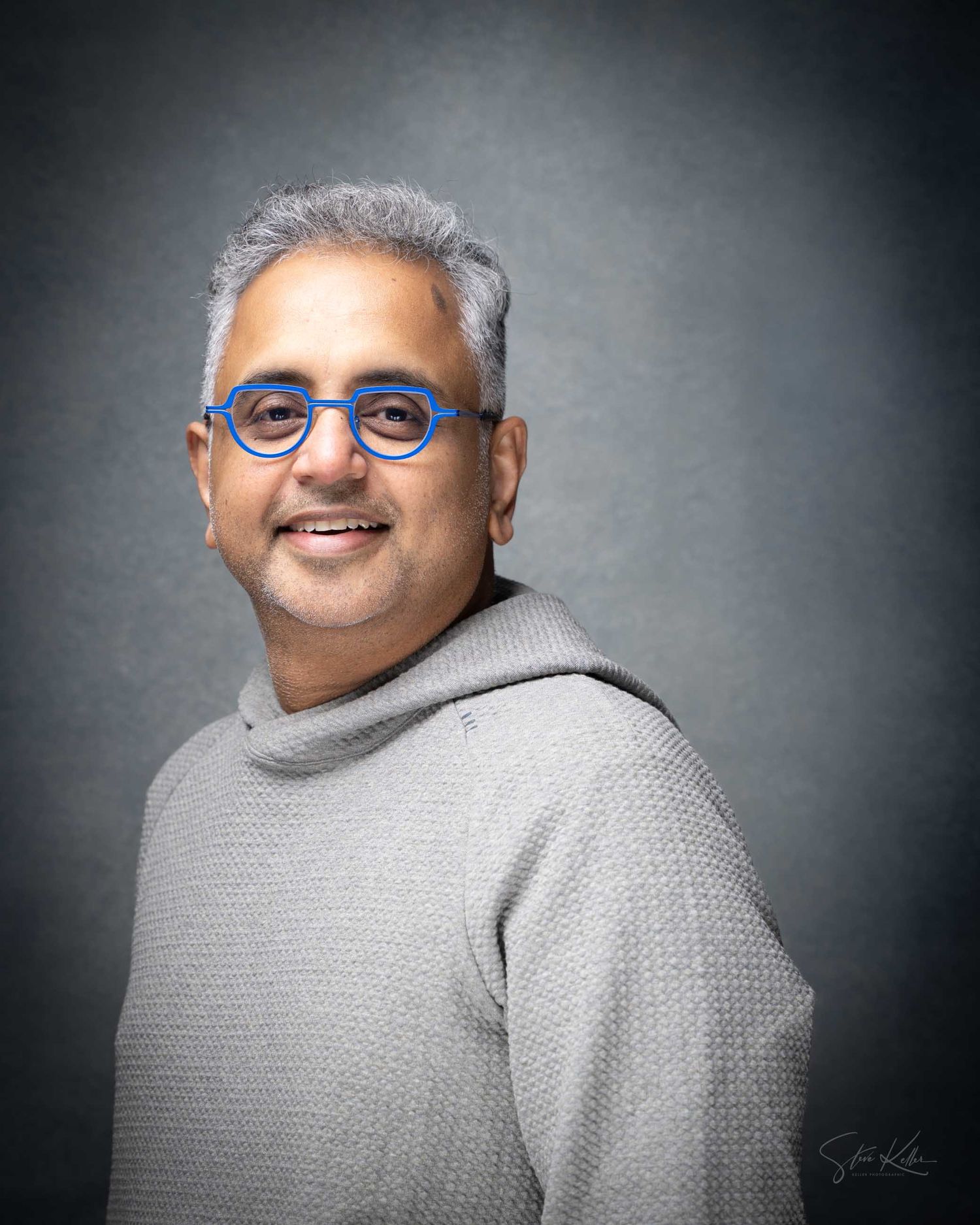 Smiling man with gray hair and blue round glasses wearing a gray hoodie sweater, professional portrait photo.