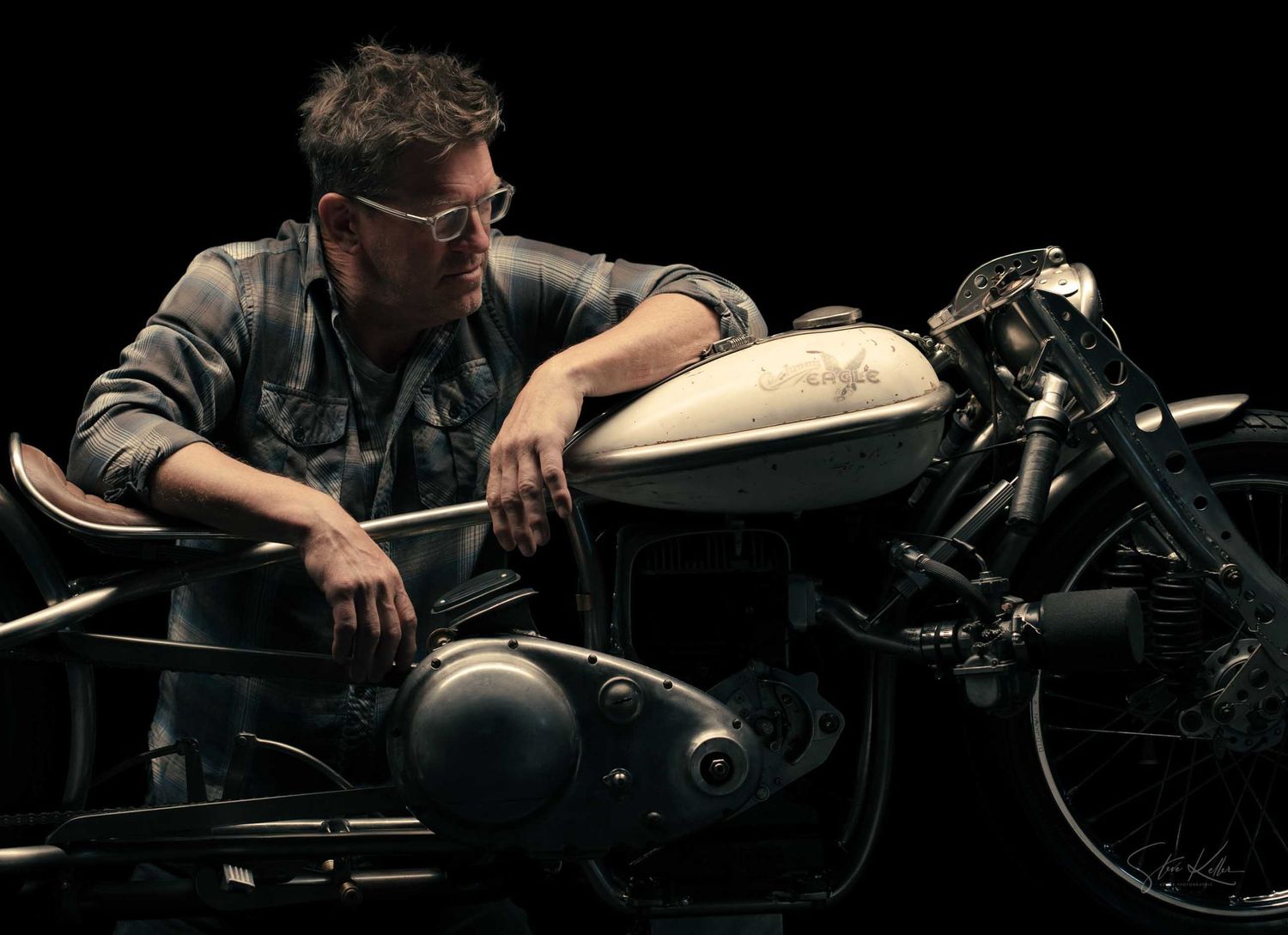 Man in denim jacket and glasses leaning on a vintage motorcycle against a dark background.