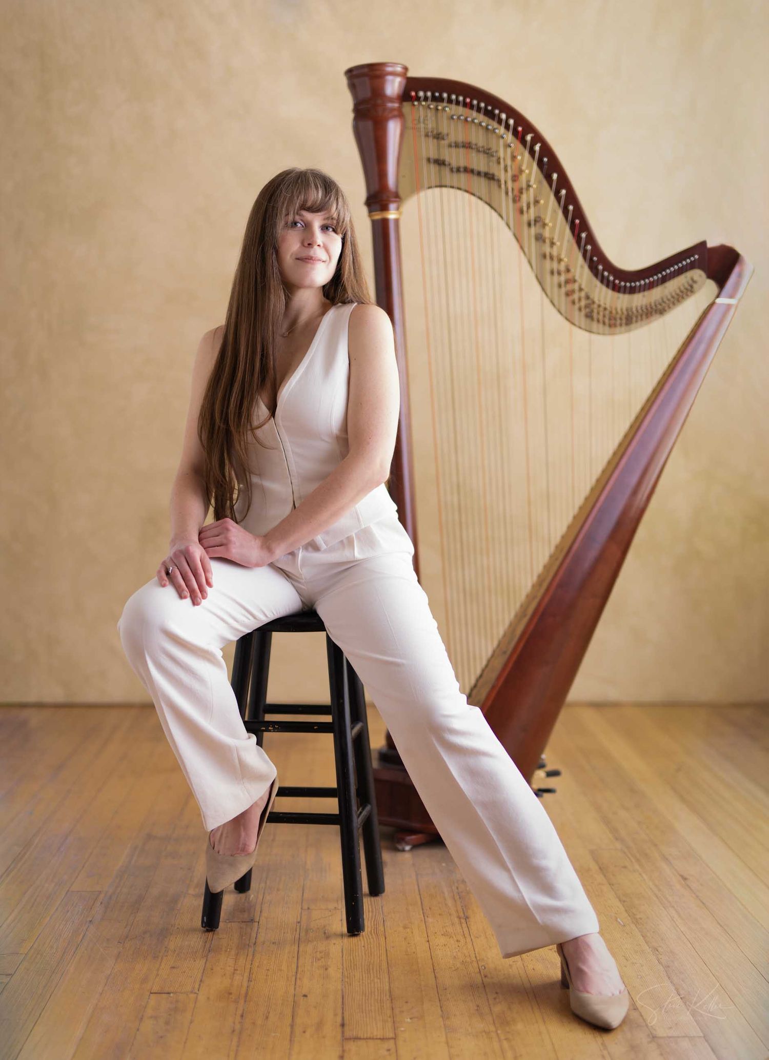 Female harpist in white outfit seated on stool beside a mahogany concert harp on a wooden floor.