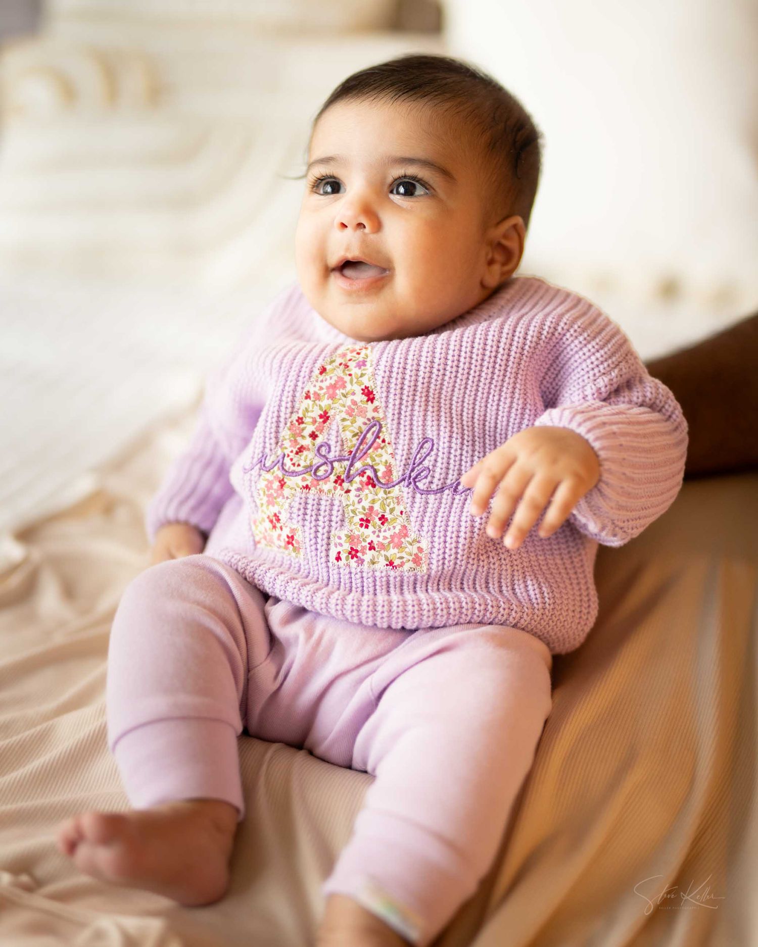 Smiling baby in lavender knit sweater and pink pants sitting upright on a bed.