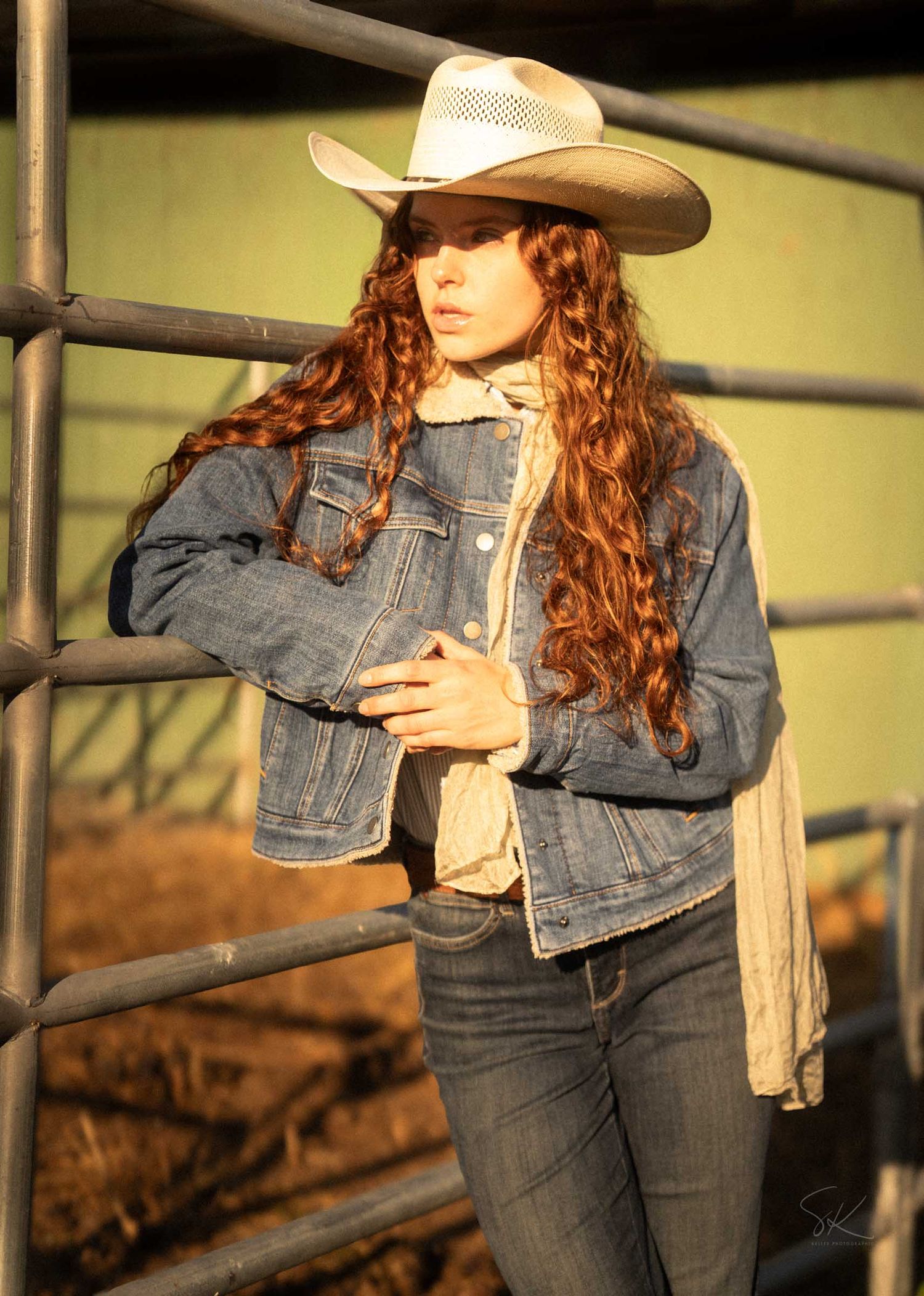 Young woman with long curly red hair wearing a denim jacket and white cowboy hat leaning on a metal fence outdoors.