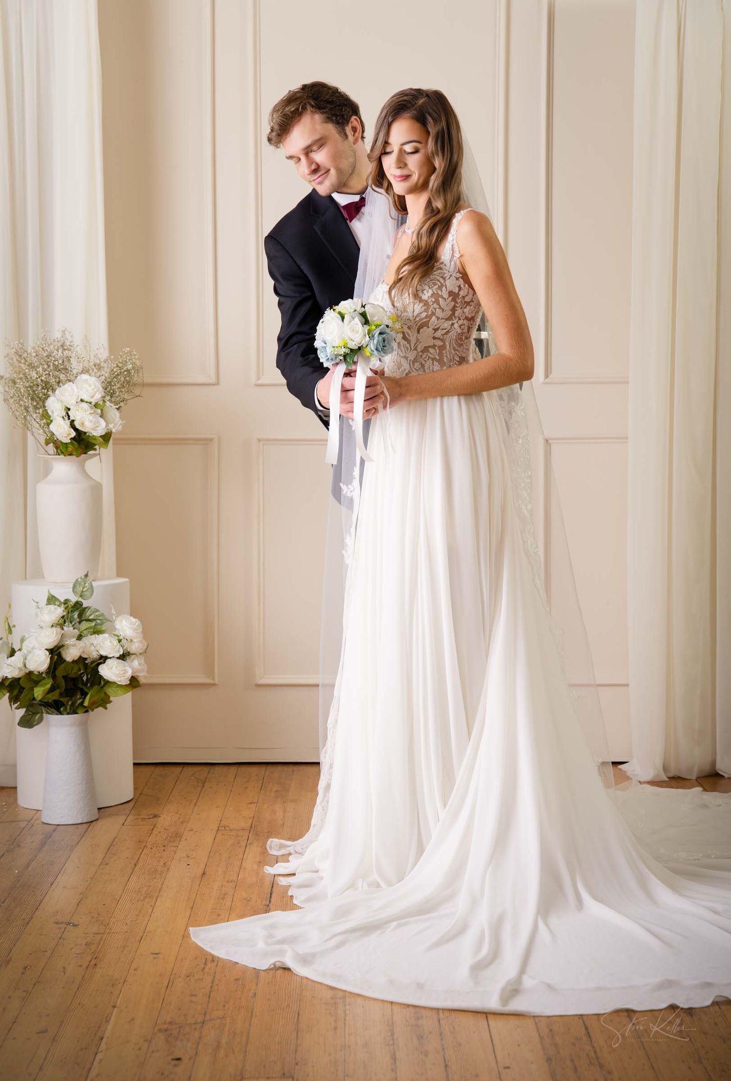Romantic wedding photo featuring couple in formal attire with white floral bouquet and long flowing bridal gown.