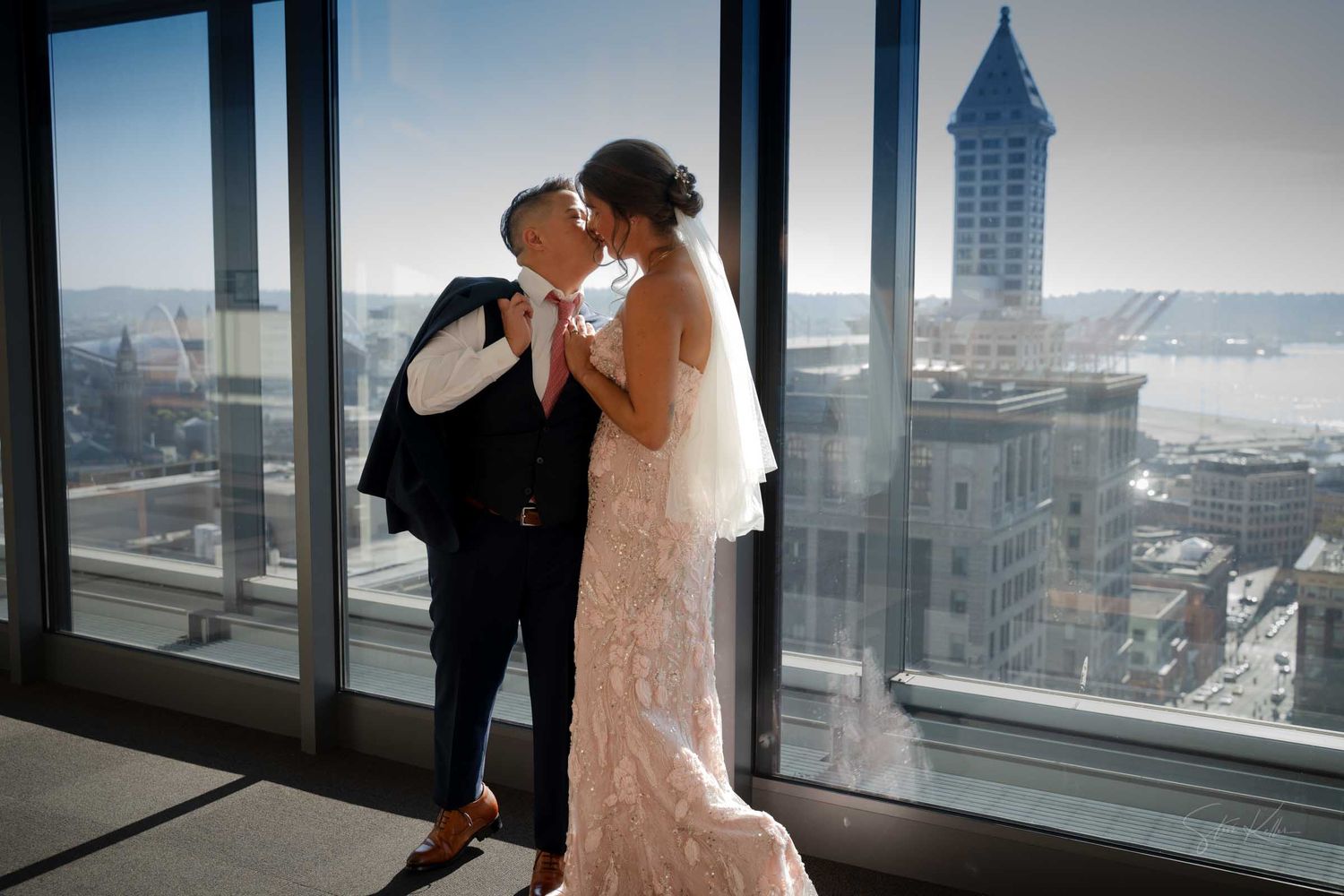 Bride and groom share a romantic kiss by floor-to-ceiling windows with a stunning Seattle skyline view.