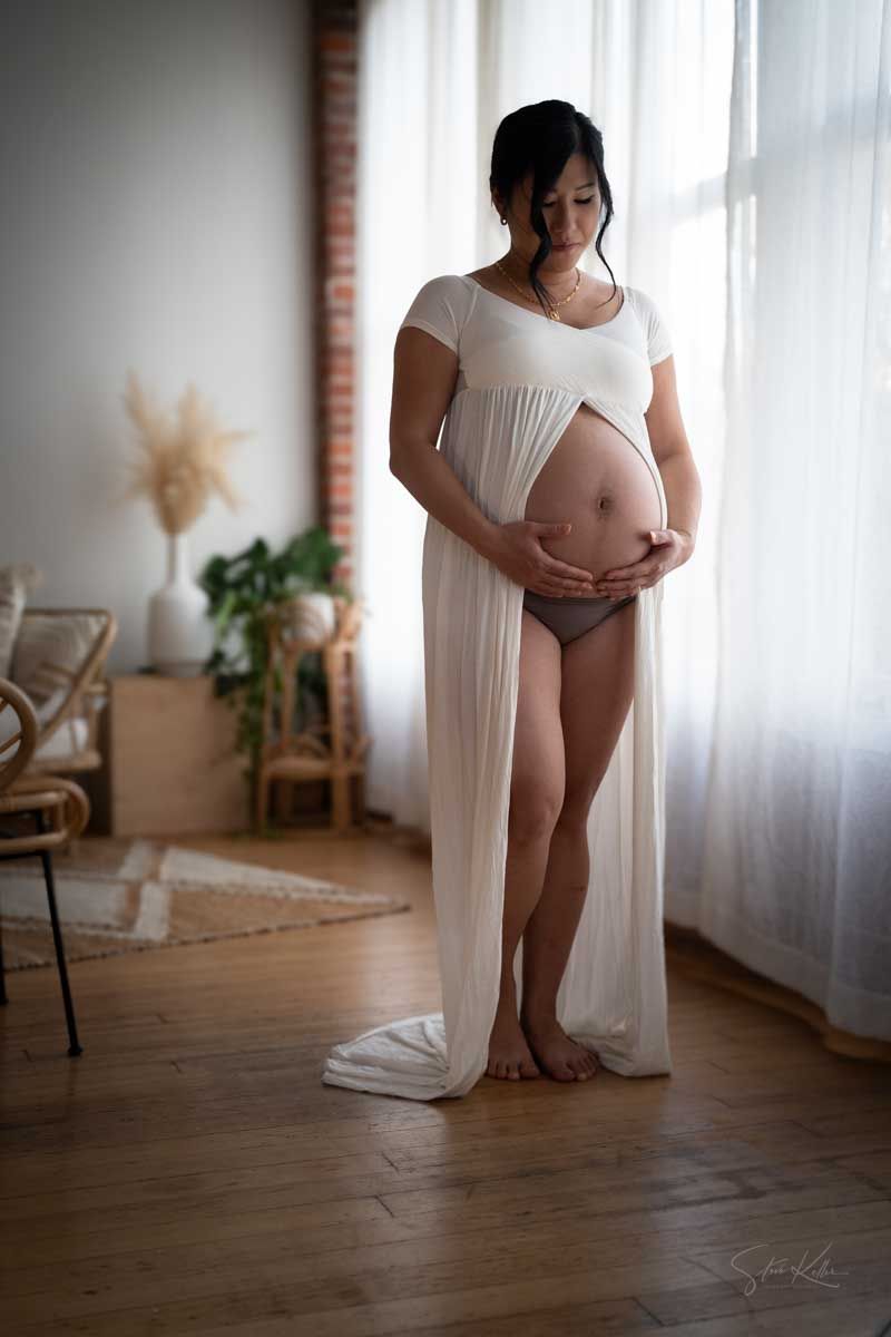 Expecting mother in a flowing white sheer gown cradles her bare belly in a bright loft studio with brick walls and wood floors.