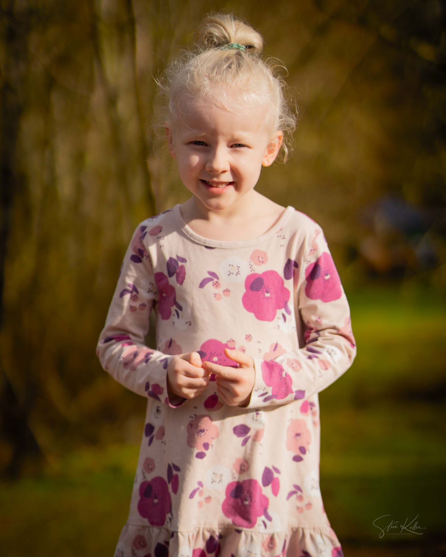 A child wearing a pink floral dress stands in a natural outdoor setting during golden hour lighting.