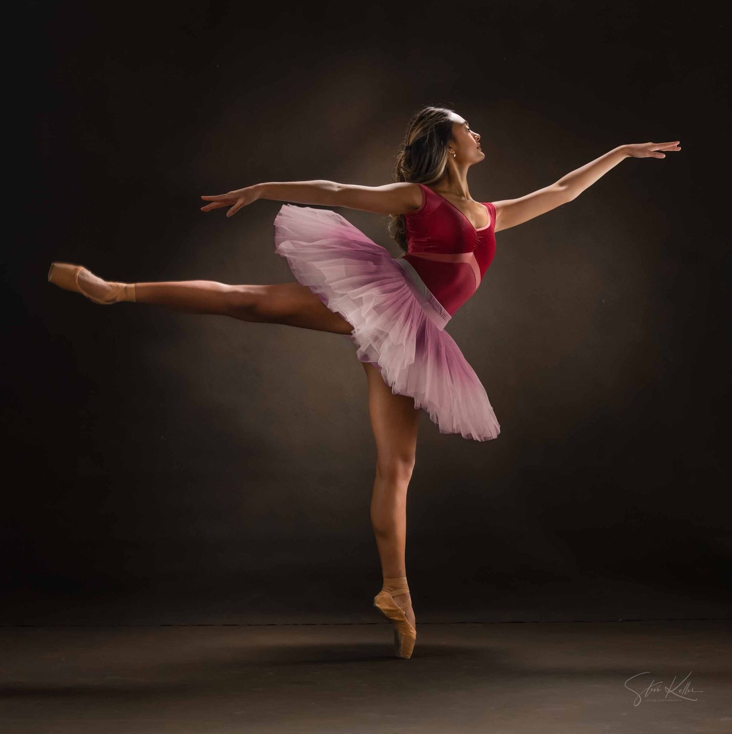 Ballet dancer in pink tutu performs graceful arabesque pose against dark background in dramatic studio lighting.