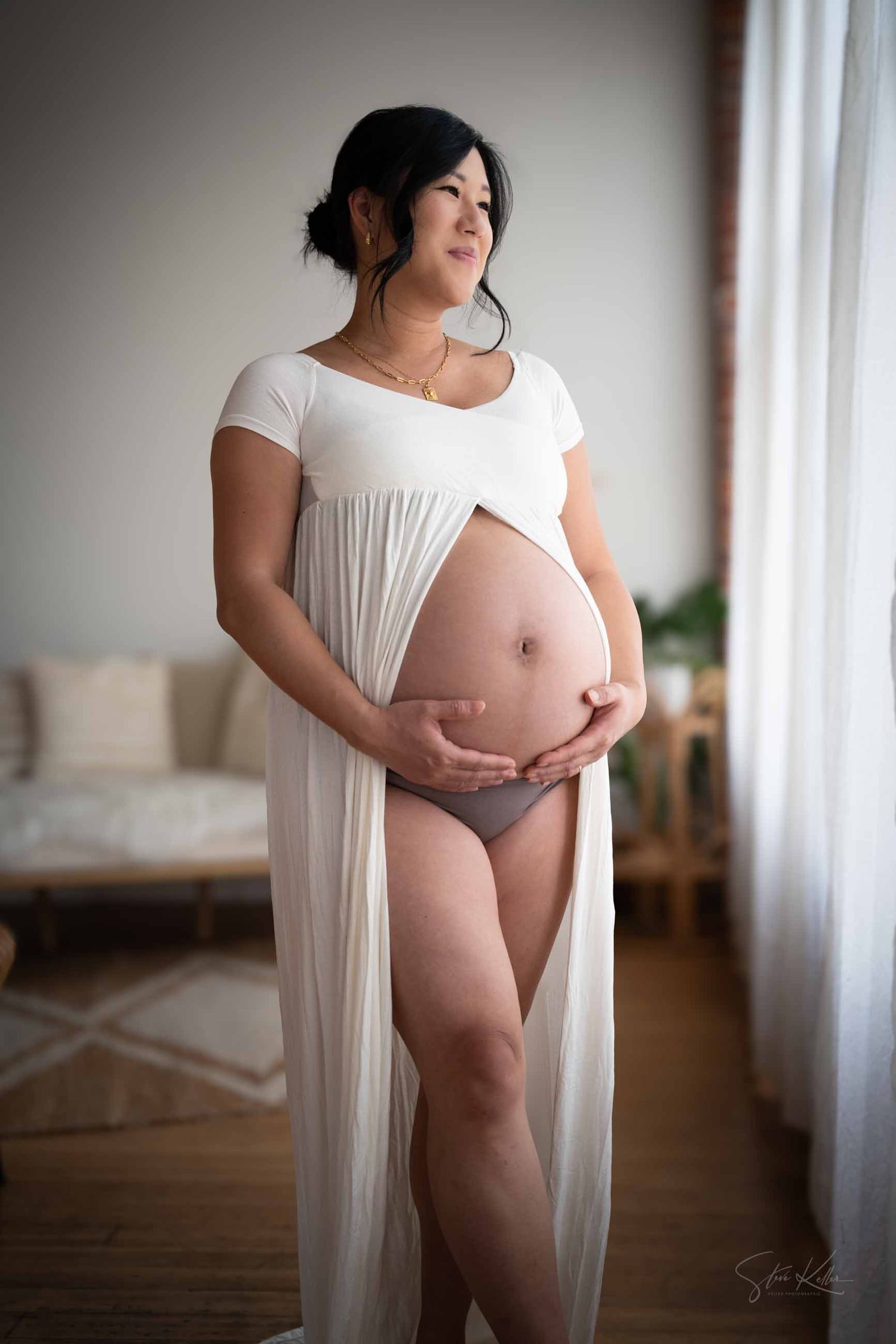 Maternity photo of an expectant mother in a flowing white dress standing by a window in soft natural light.