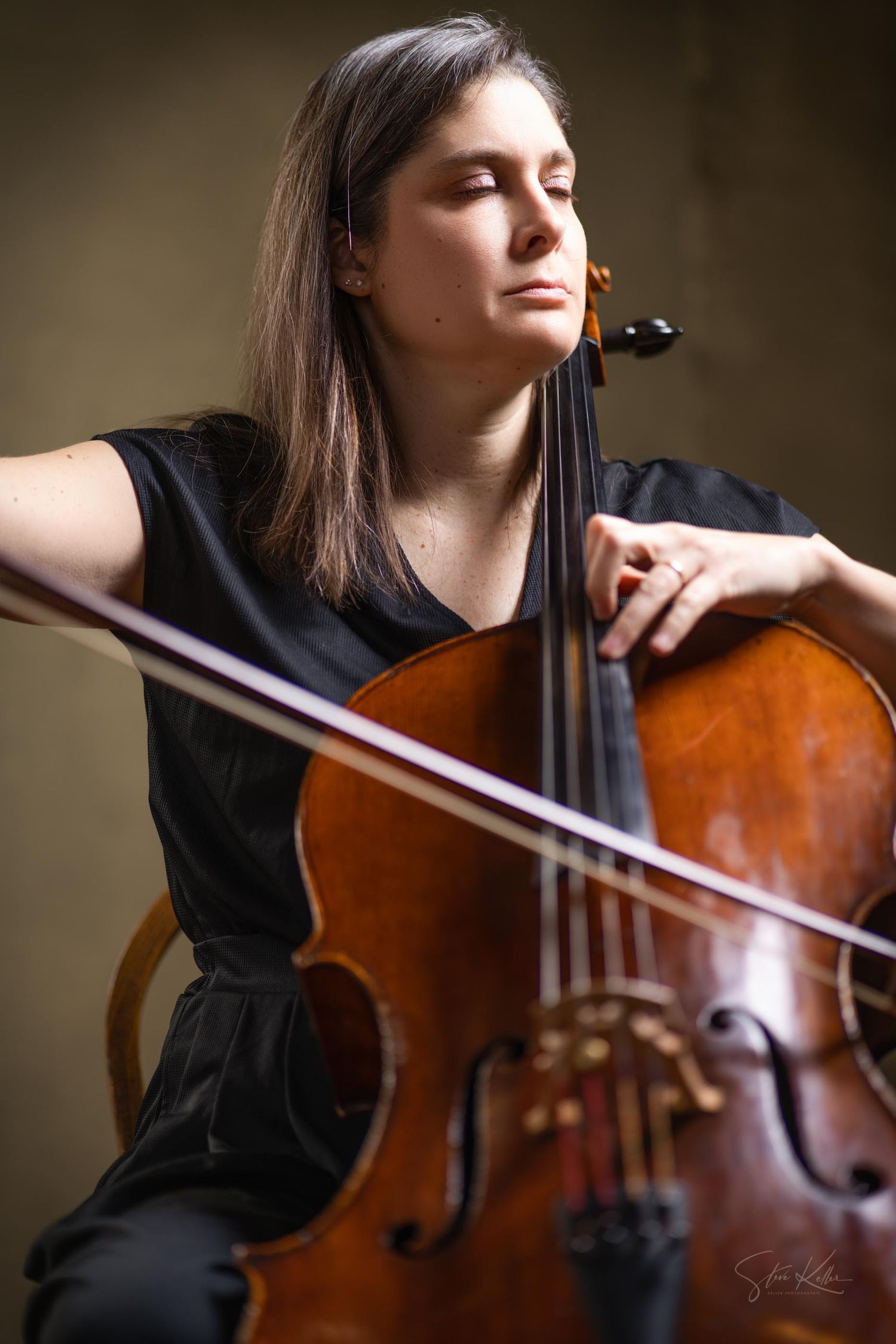 A cellist in black attire performs with their wooden instrument against a dark atmospheric background.