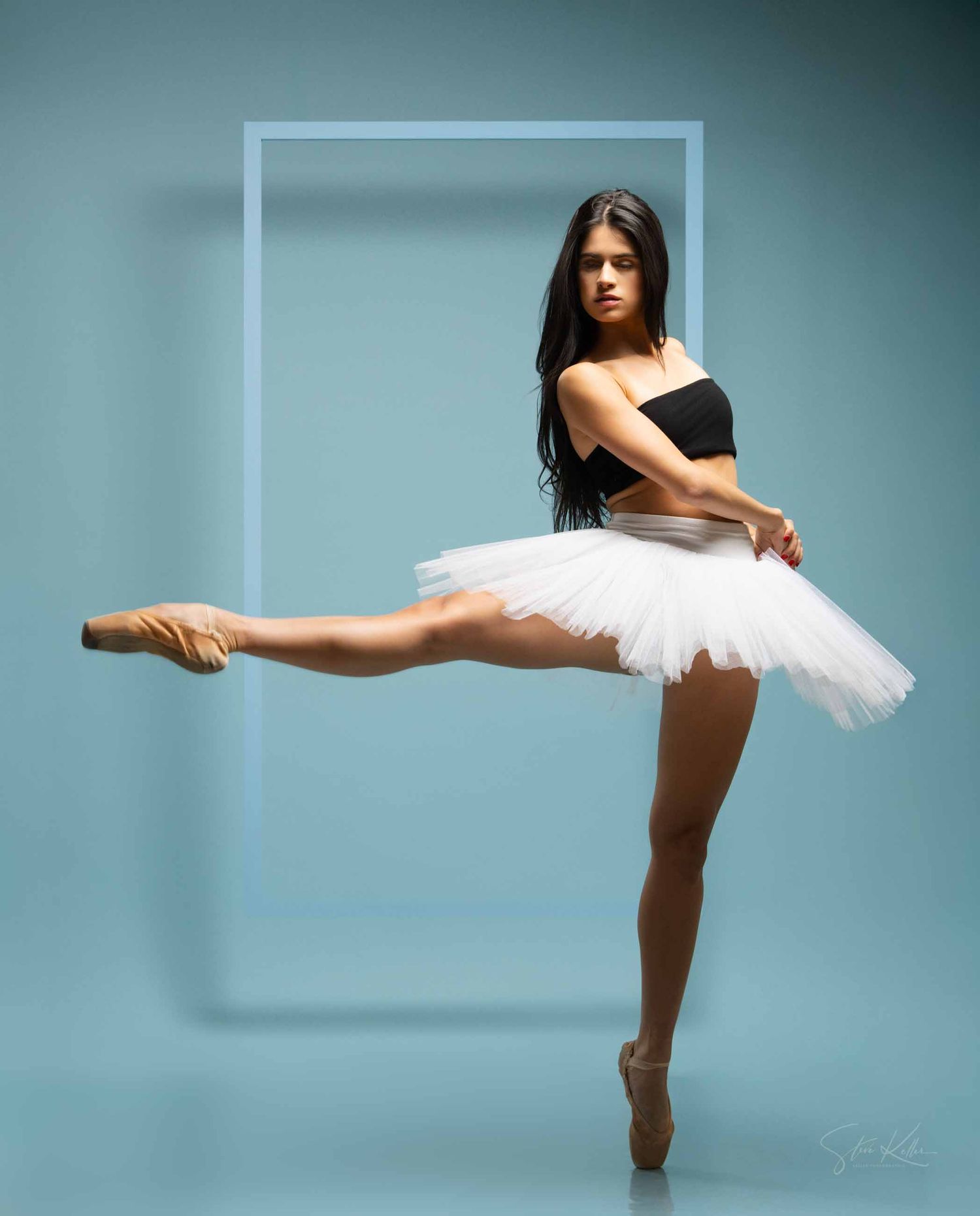 Ballerina in white tutu performs elegant arabesque pose against turquoise background with geometric frame element.