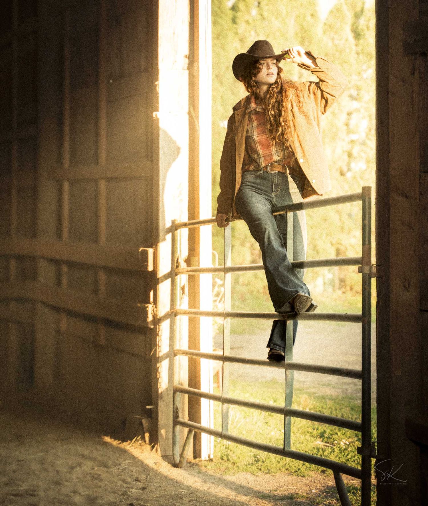 Woman in cowboy hat and jeans posing on a ranch gate in golden sunlight inside a rustic barn doorway.