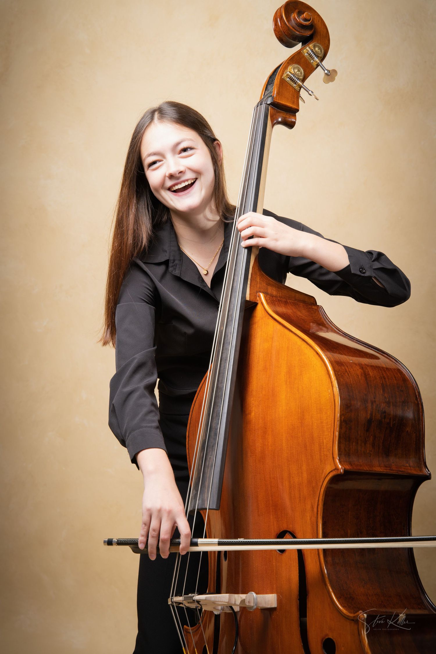 A musician in a black shirt performs with a wooden double bass against a neutral background.