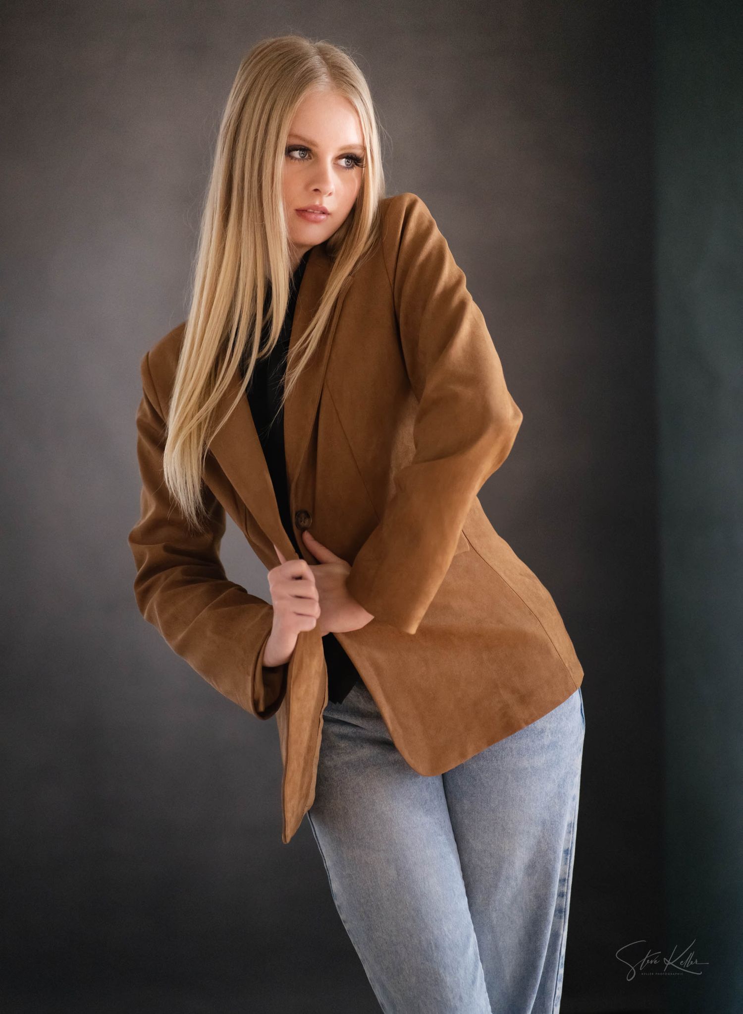 Blonde woman posing in a tan suede blazer and light blue jeans against a dark gray studio background.