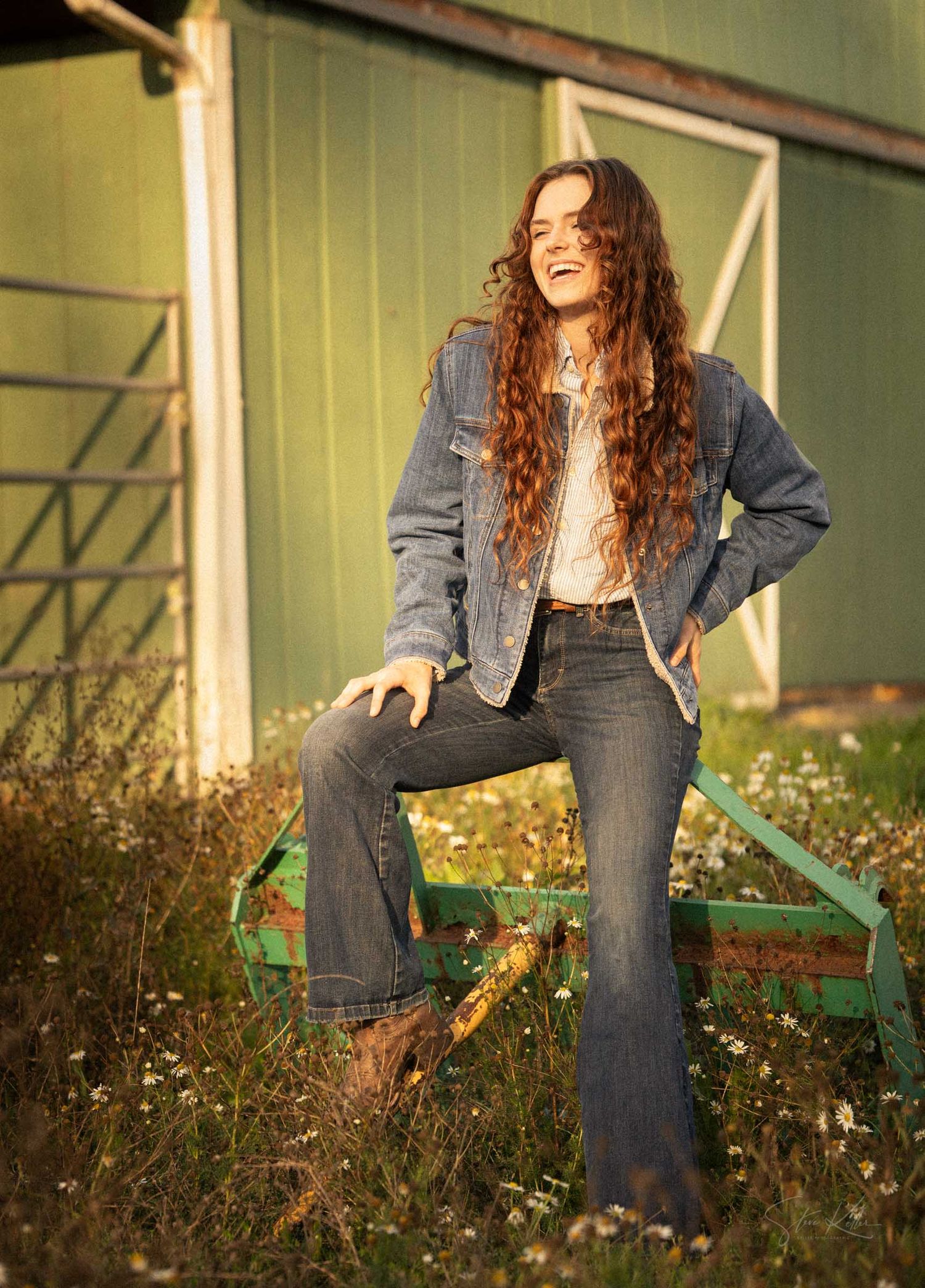 Smiling woman with long curly hair wearing denim jacket and flare jeans poses outdoors near a green barn.