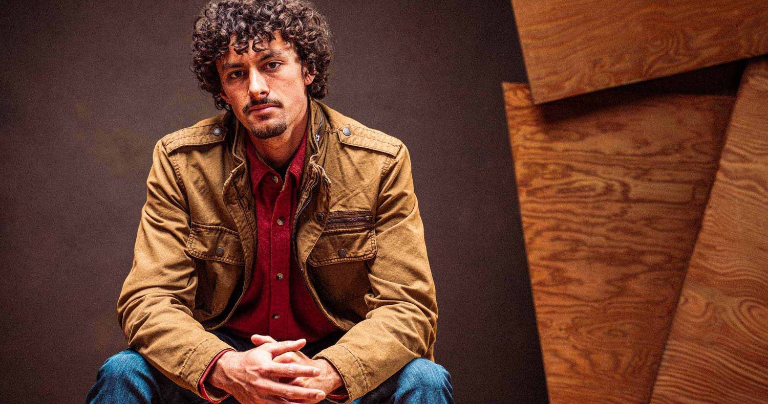 Man with curly hair in a tan work jacket and red shirt sits with clasped hands against a dark backdrop with wooden panels.