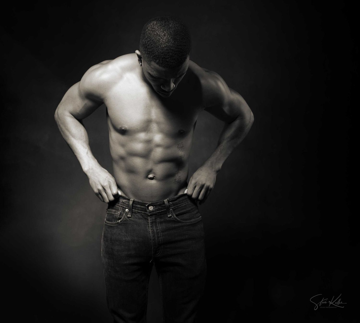 Black and white photo of a muscular shirtless man with defined abs posing with hands on hips.