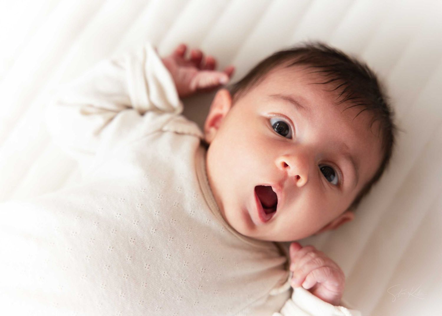 A baby lies on white bedding with an expressive surprised look while wearing a cream colored outfit.