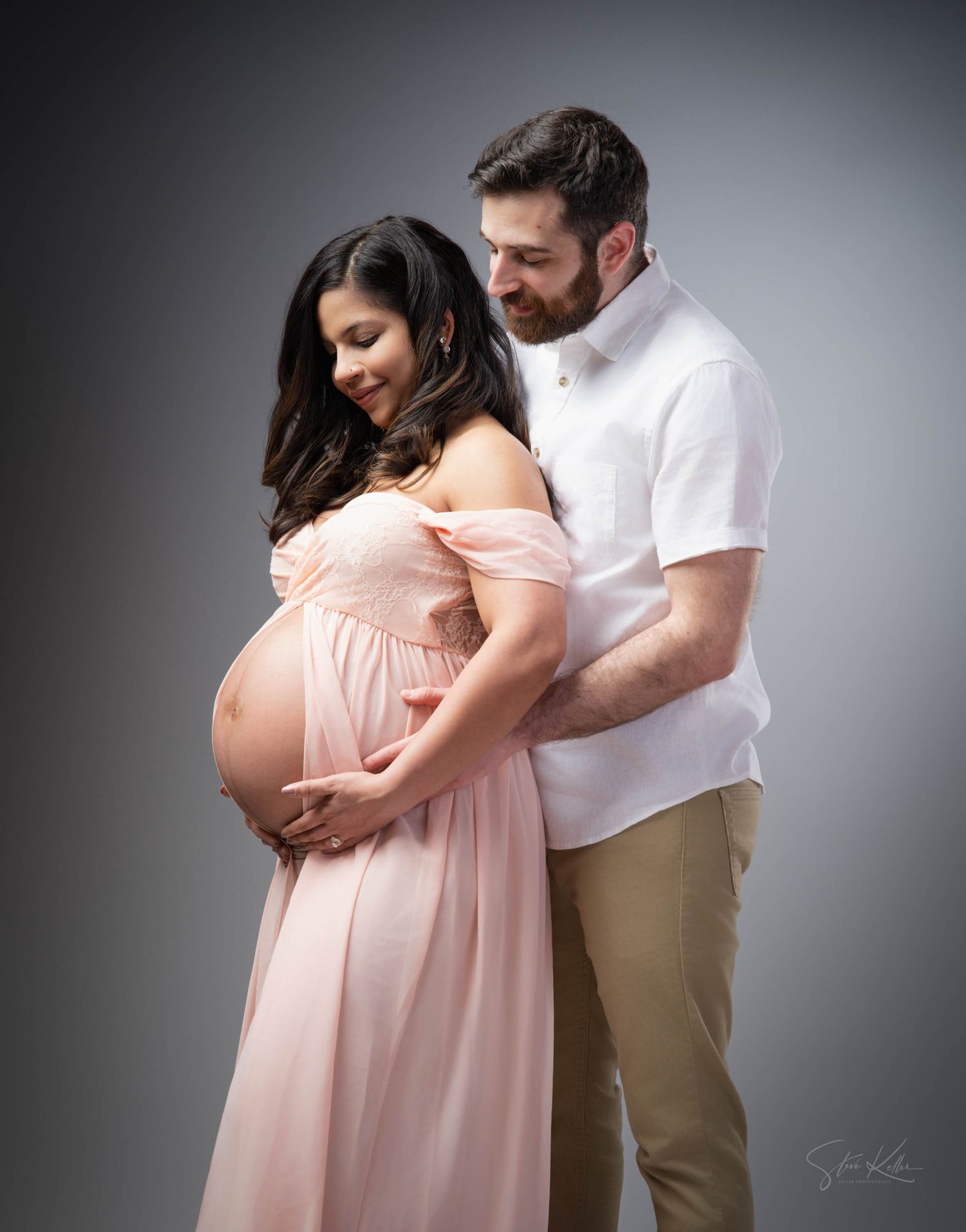 A couple in white and beige share a tender moment in a studio maternity photo shoot with a pink flowing dress.