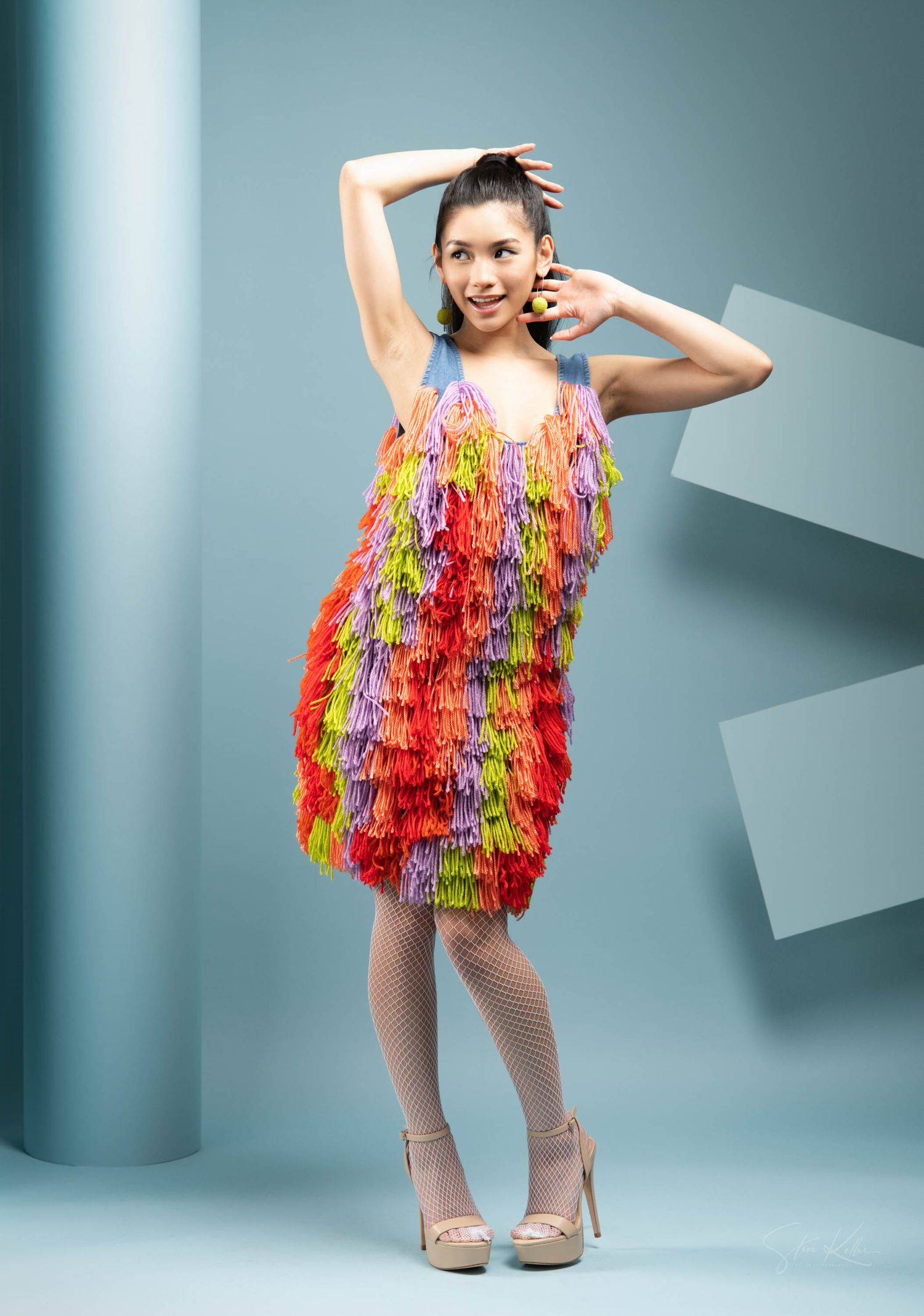 Colorful fringed party dress in rainbow hues photographed against blue studio backdrop.