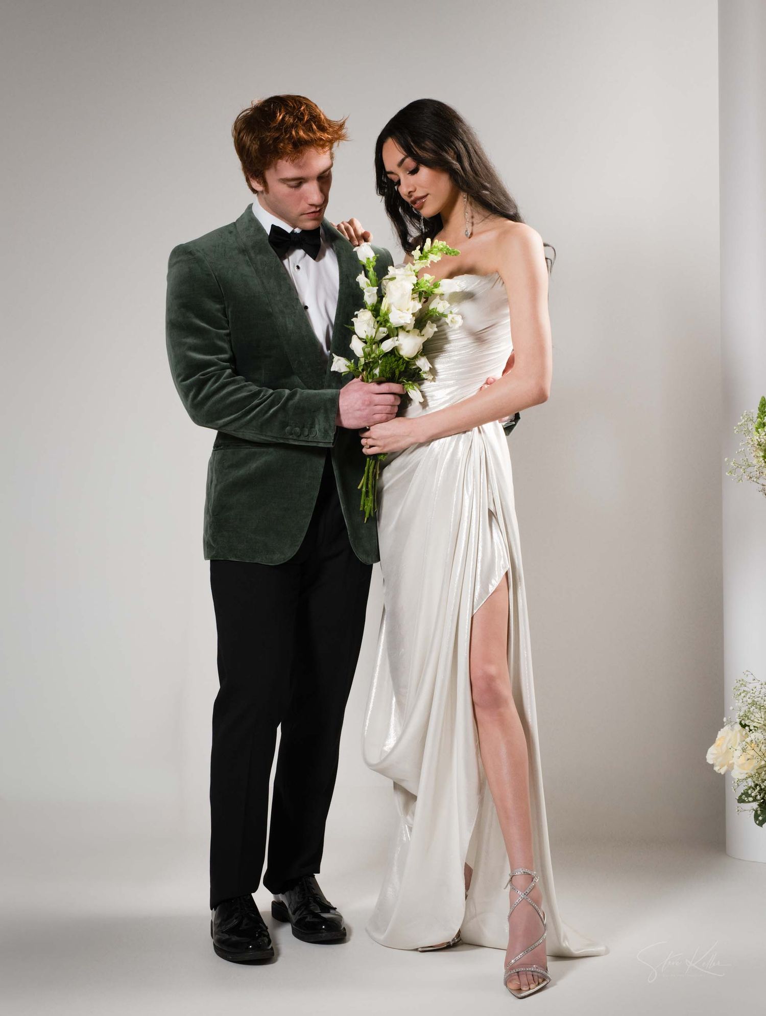 A romantic couple in formal attire poses together against a white studio backdrop with elegant white flowers.