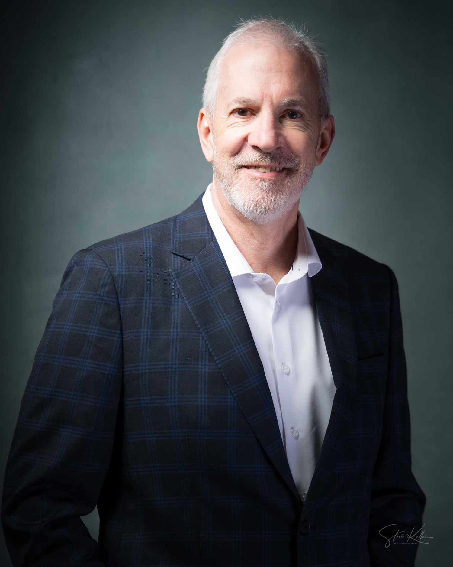 Professional headshot of a smiling middle-aged man with white hair and beard wearing a plaid blazer.