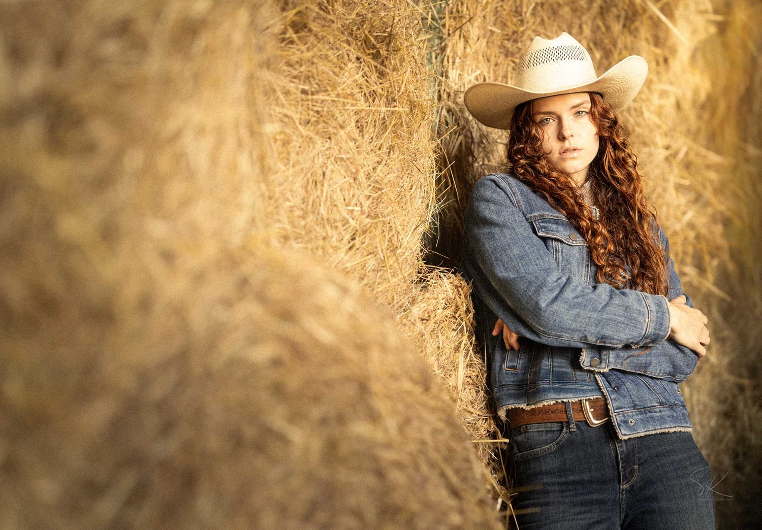Young woman in cowboy hat and denim jacket leaning against hay bales on a farm.