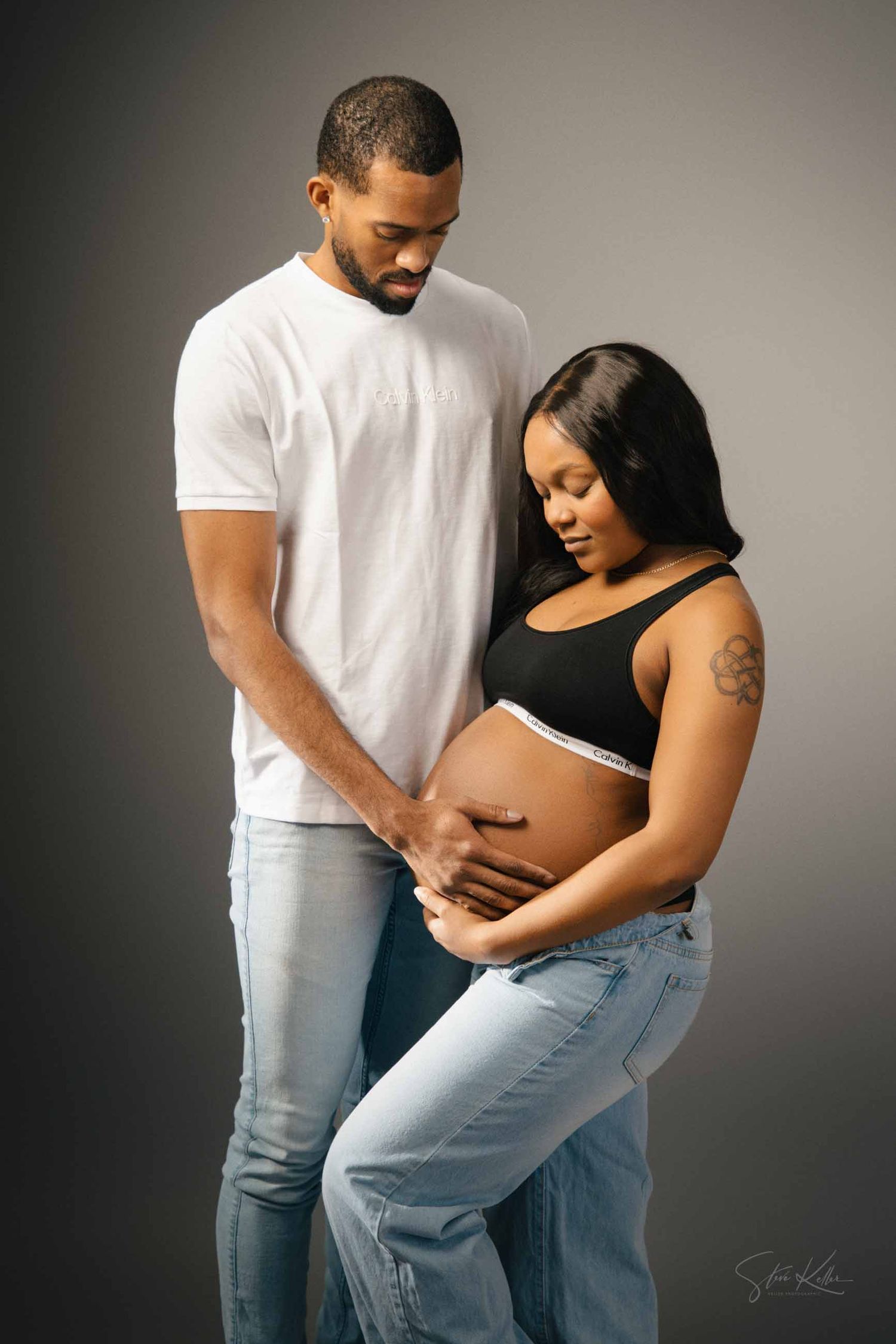 A couple in casual attire share an intimate moment during a maternity photoshoot against a gray backdrop.