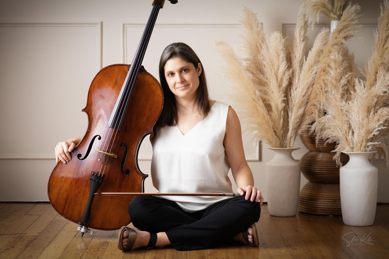 A cellist performs with their instrument in a studio setting with pampas grass decor and natural lighting.