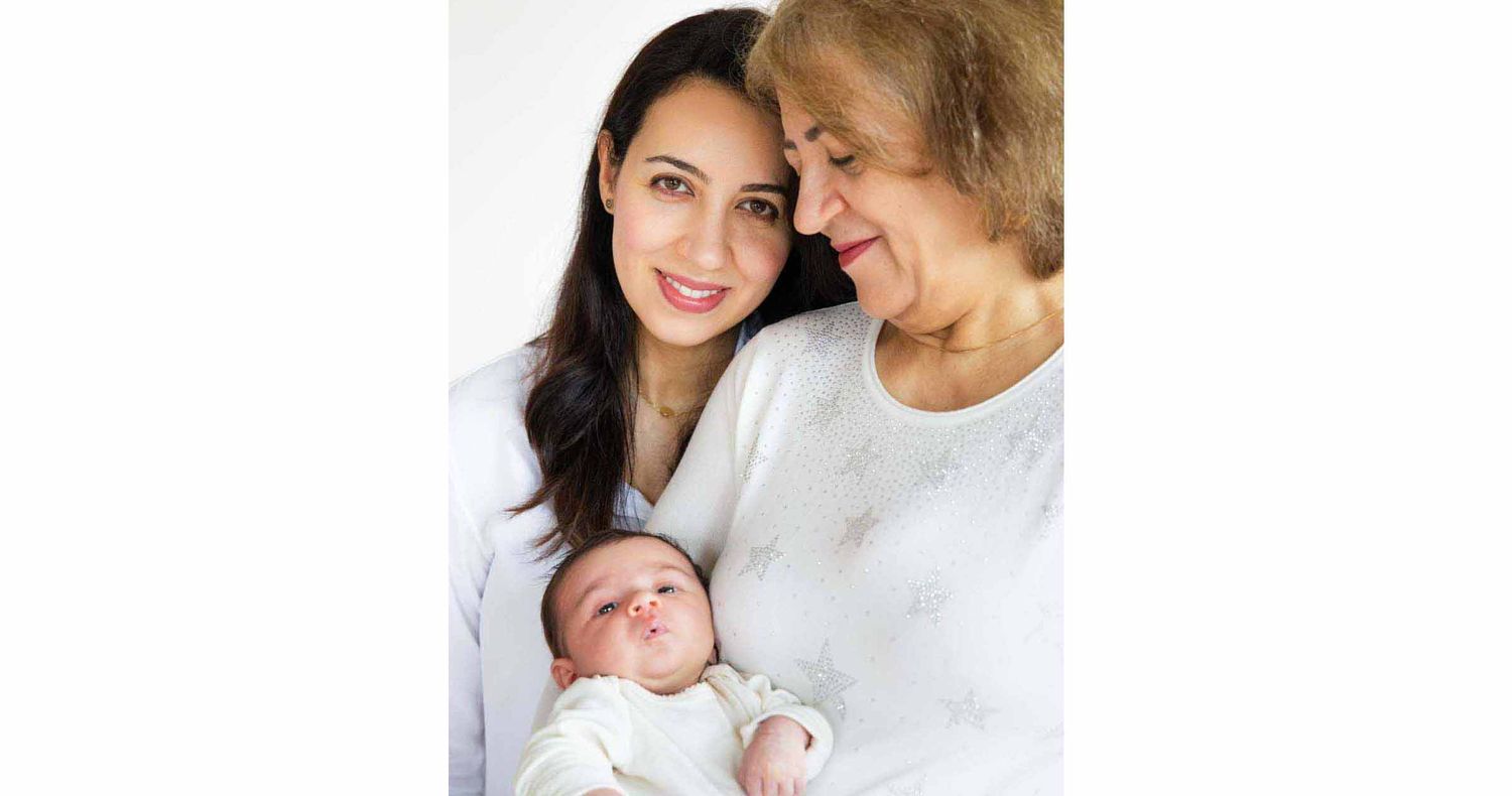 A smiling young woman and an older woman in white tops lovingly pose together while holding a sleeping newborn baby.