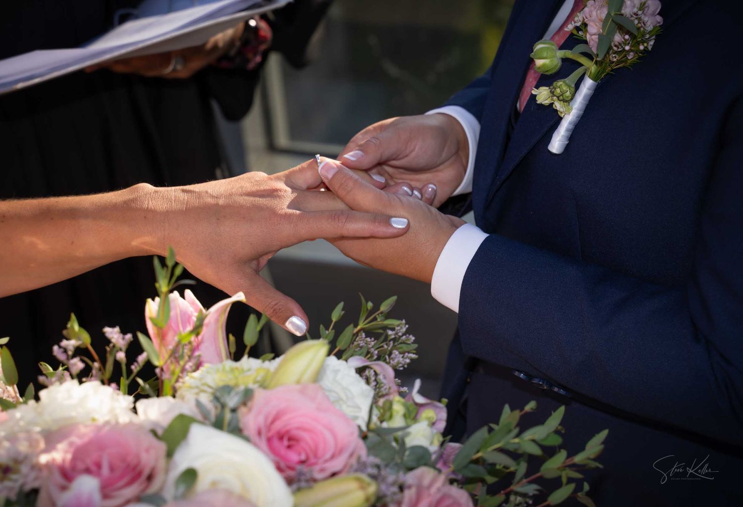 Close-up of groom placing wedding ring on brides finger over pink floral bouquet.