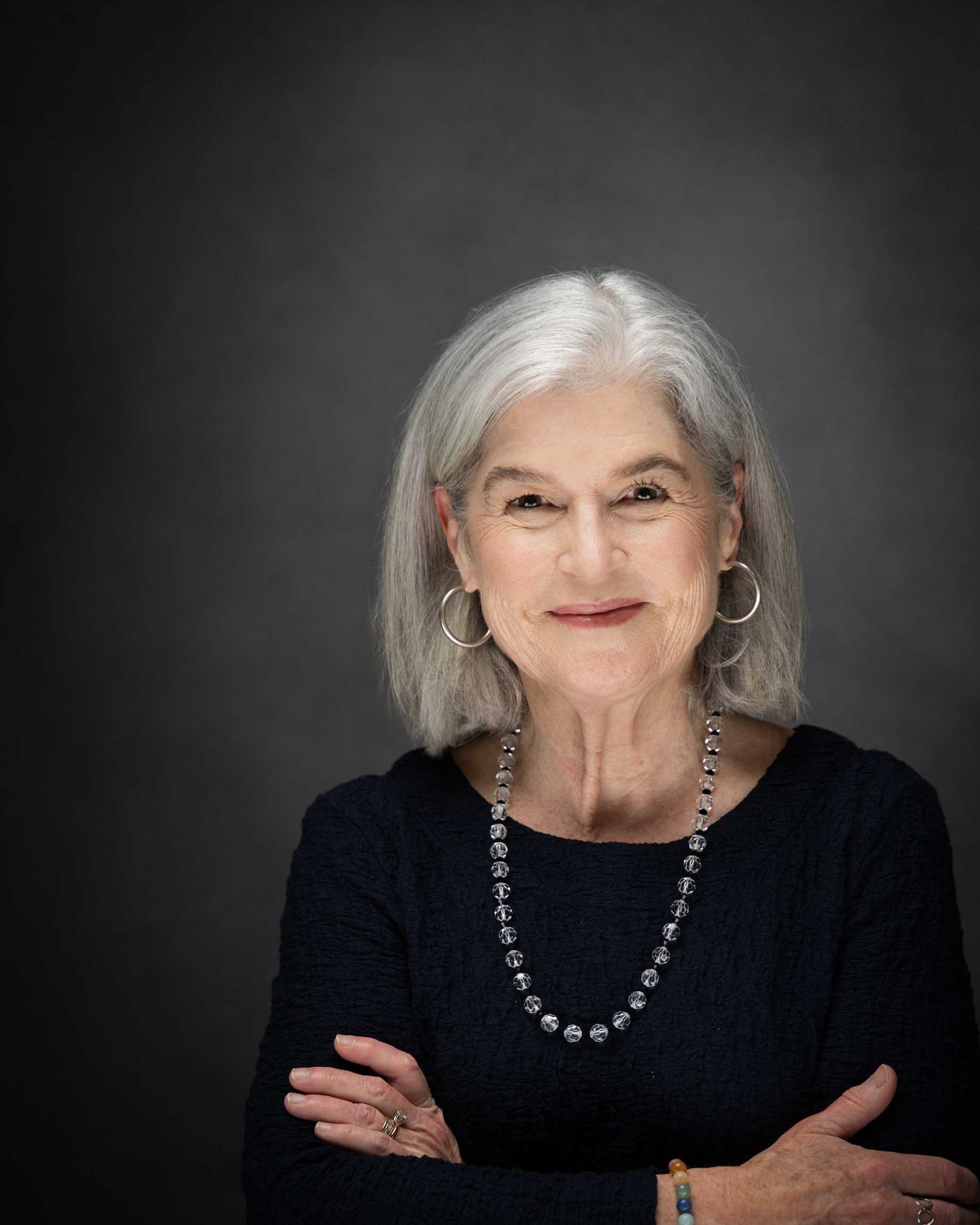 Elegant older woman with silver hair, hoop earrings, beaded necklace, and crossed arms, smiling against gray background.