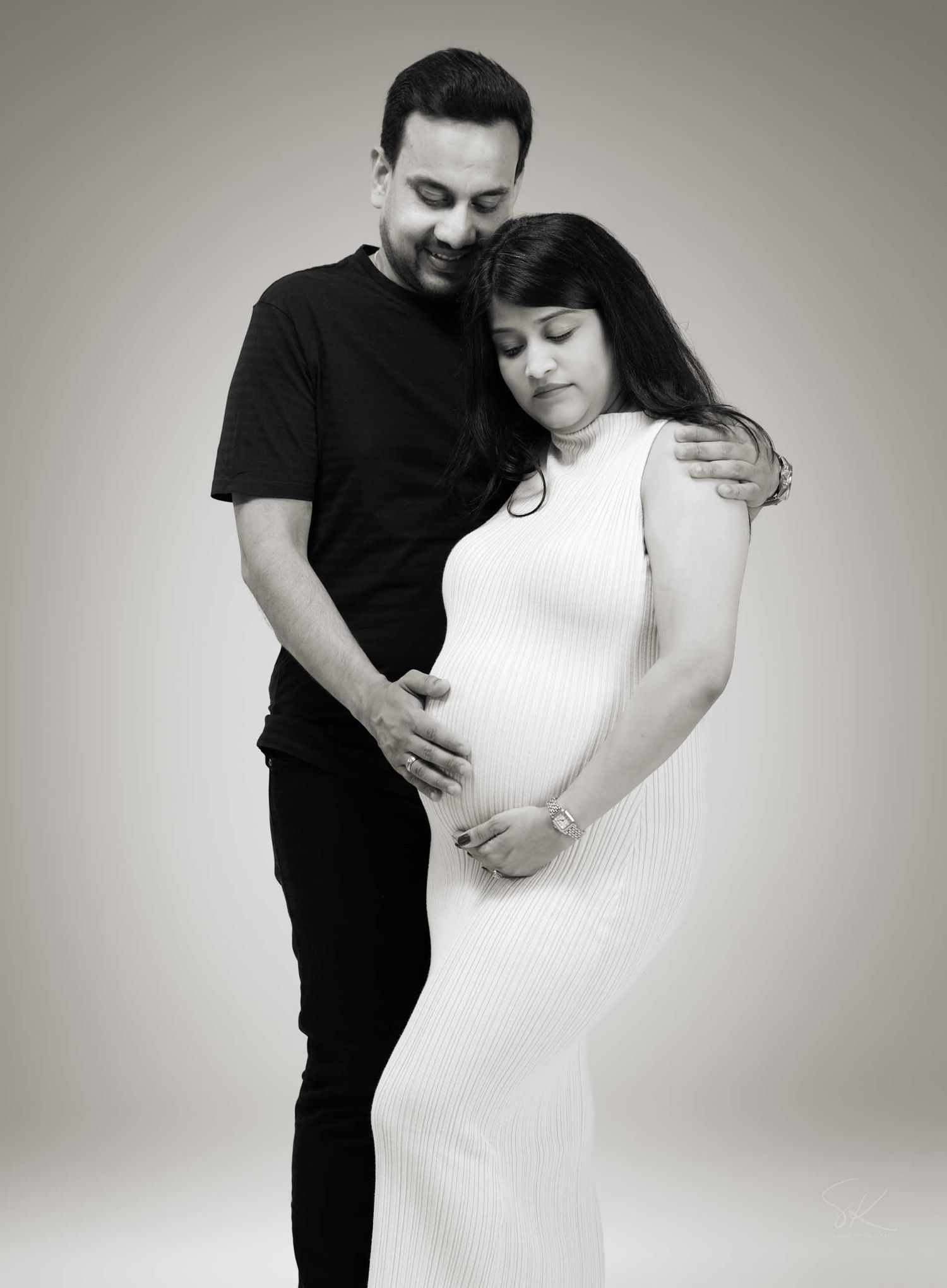 Black and white maternity photo of a couple, man in black embracing pregnant woman in white ribbed dress.