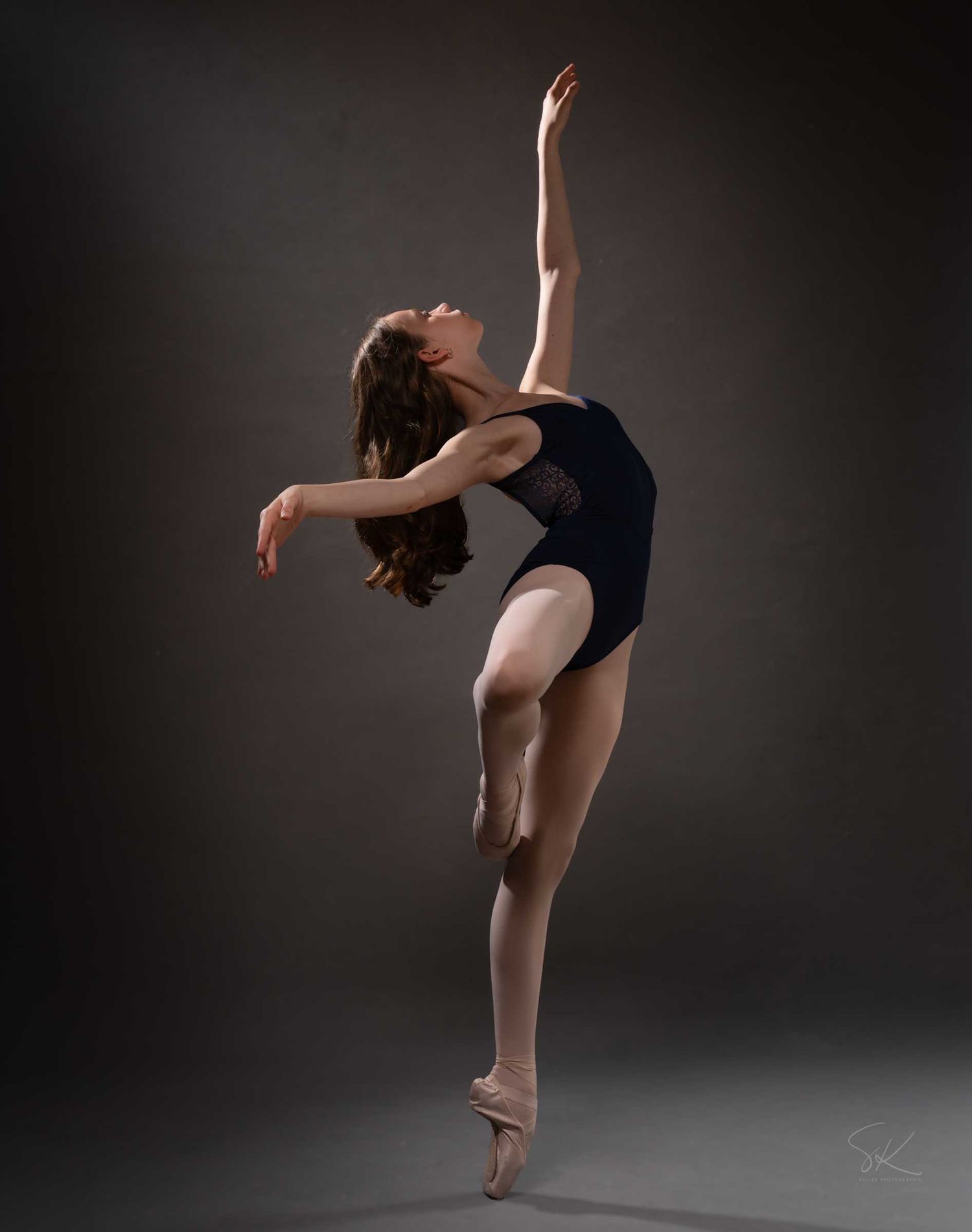A ballet dancer in a navy leotard and pointe shoes performs an elegant arabesque pose against a dark gray background.