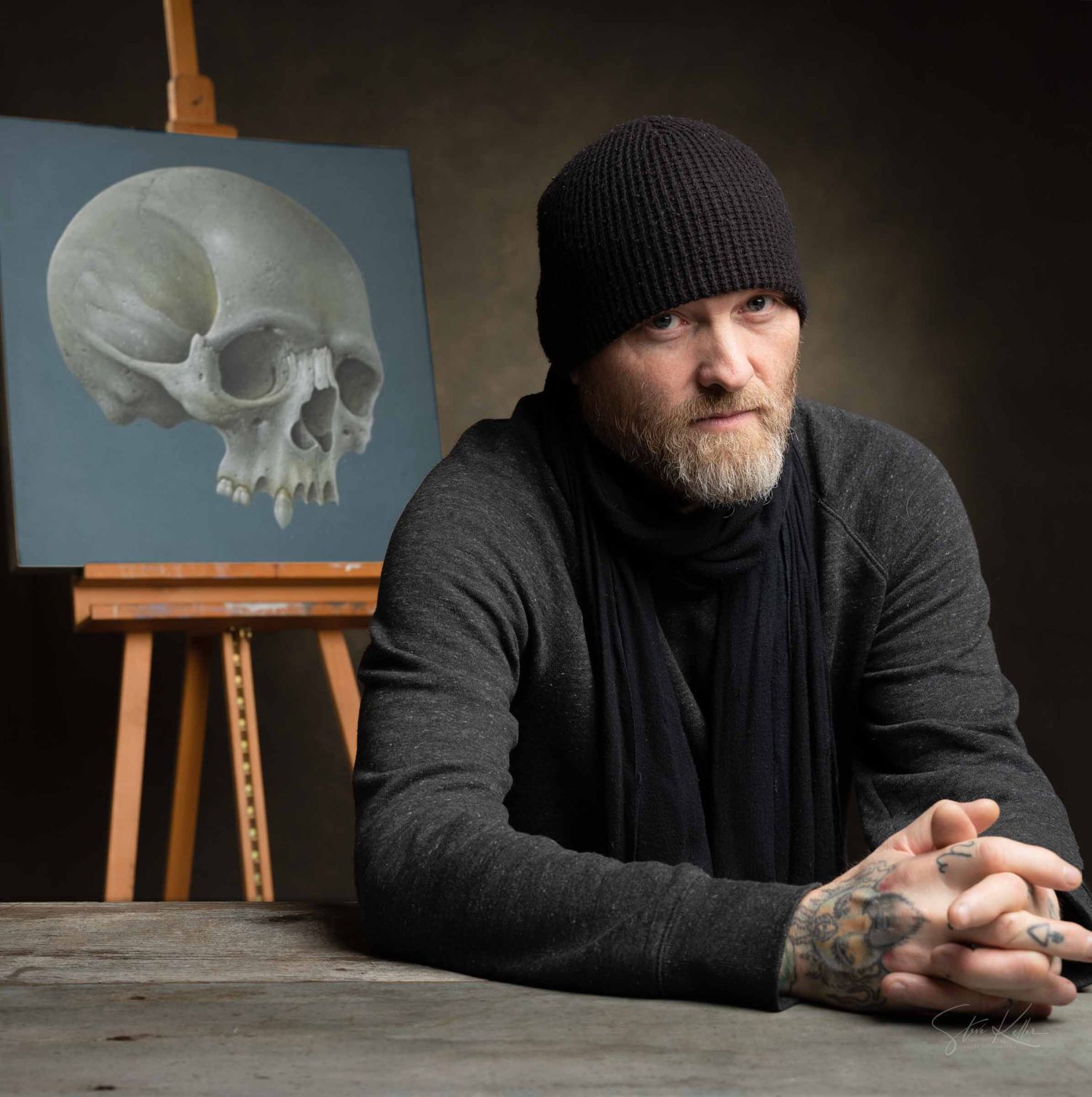 Artist in dark attire sits at wooden table with skull sculpture and easel in moody studio setting.