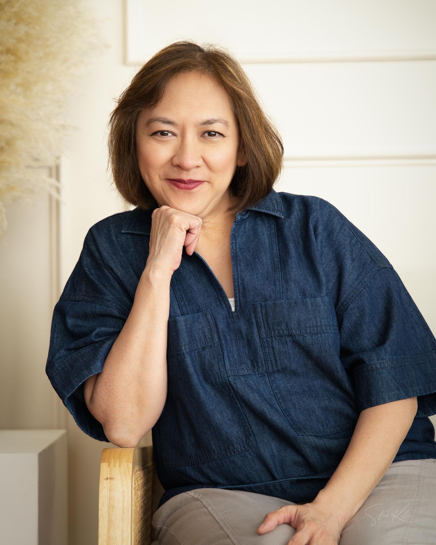 A person in a denim shirt is resting their chin on their hand while sitting in a professional headshot style portrait session.