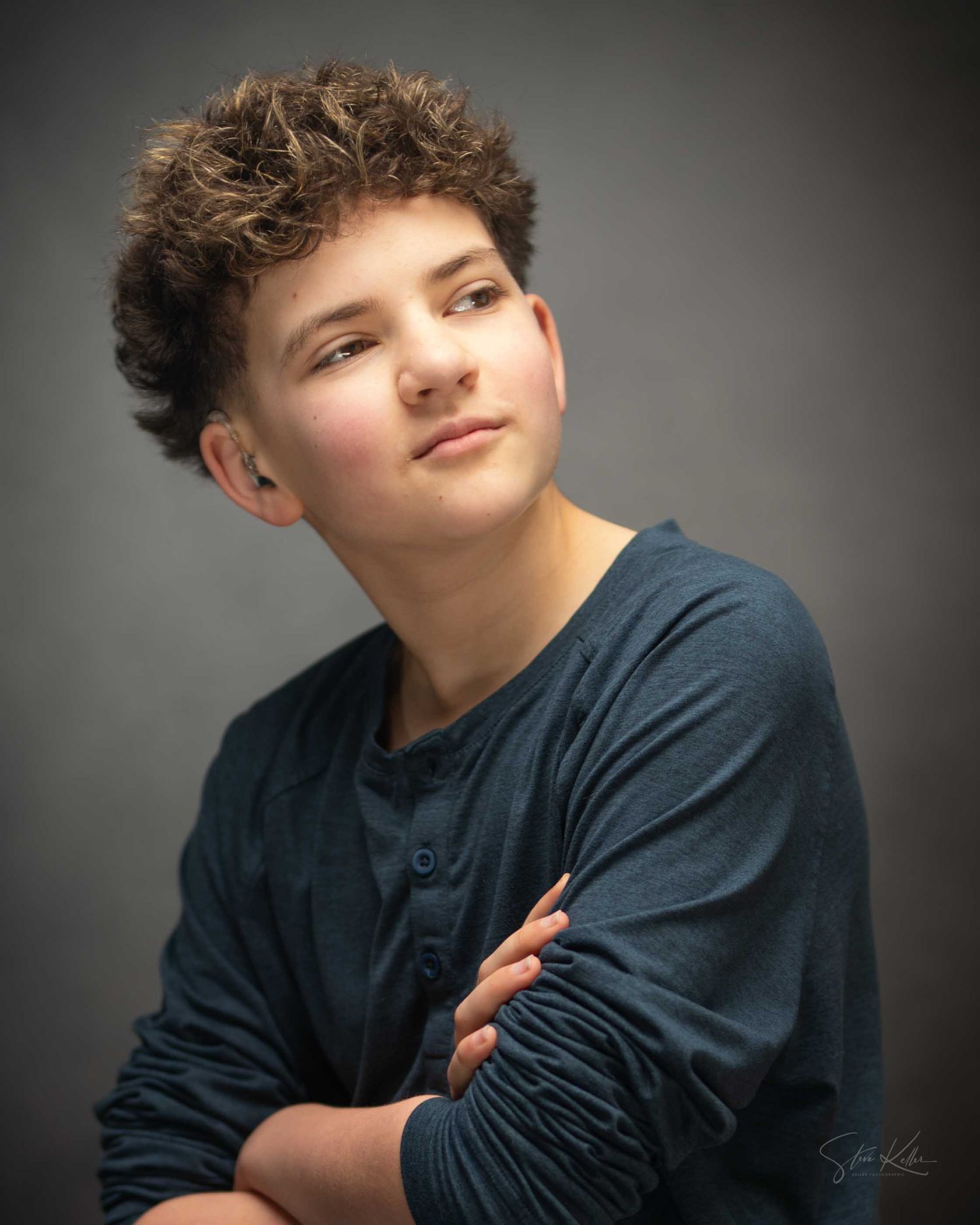 A young teen boy with curly brown hair poses with arms crossed in a blue henley shirt against a gray studio background.