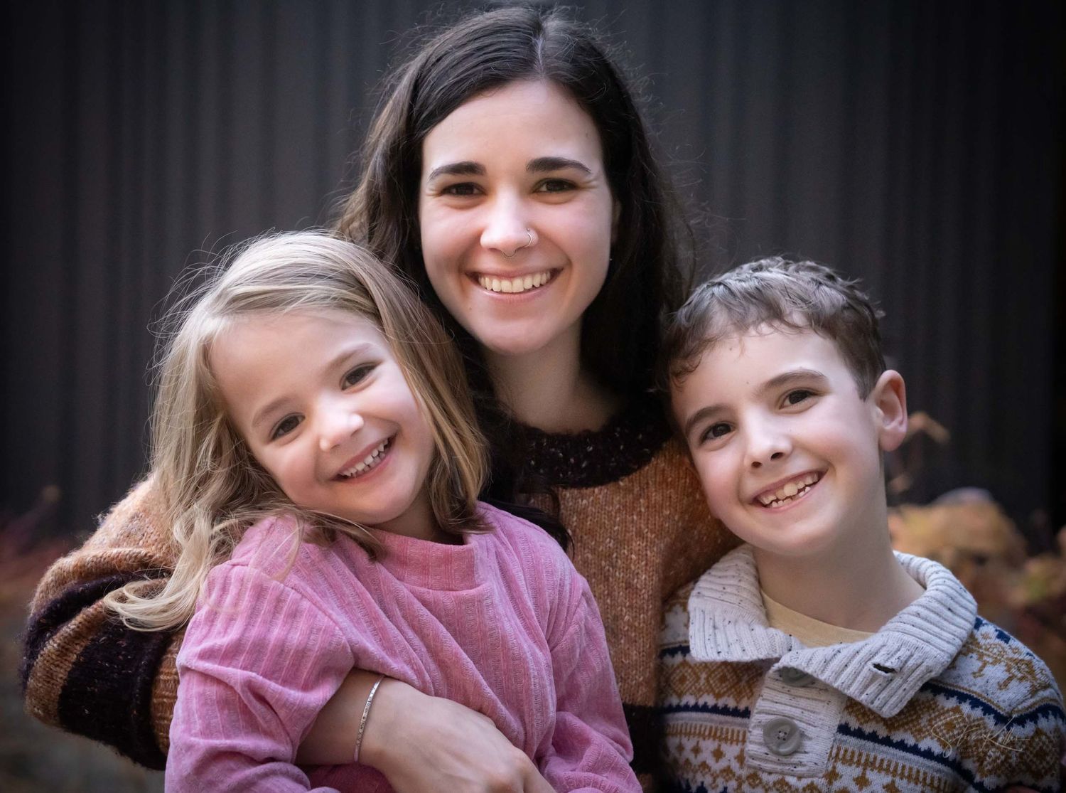 A smiling woman poses with two happy children in a warm family portrait outdoors.