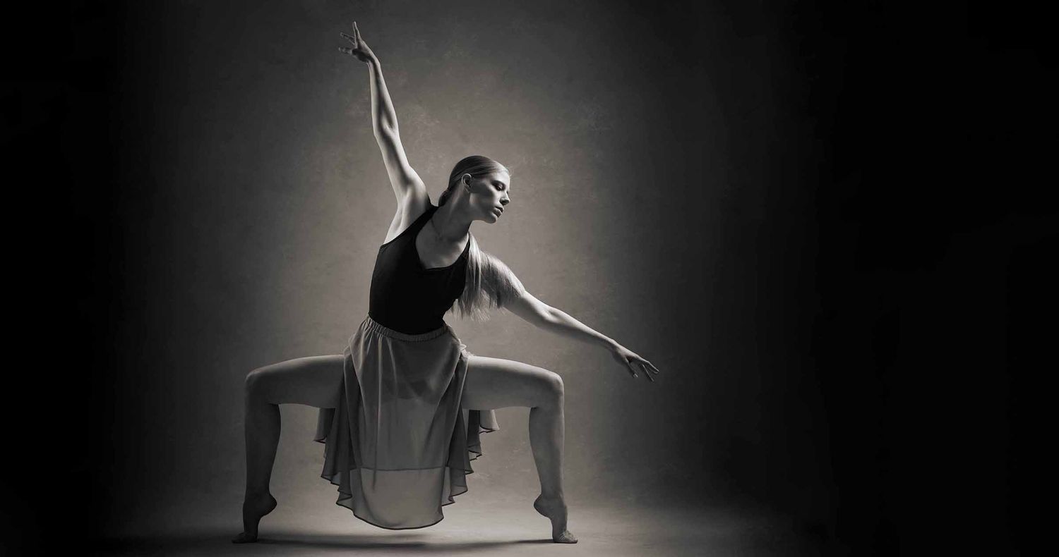 Female contemporary dancer in a wide plié pose with arms extended gracefully, wearing a flowing skirt against a dark moody studio backdrop.