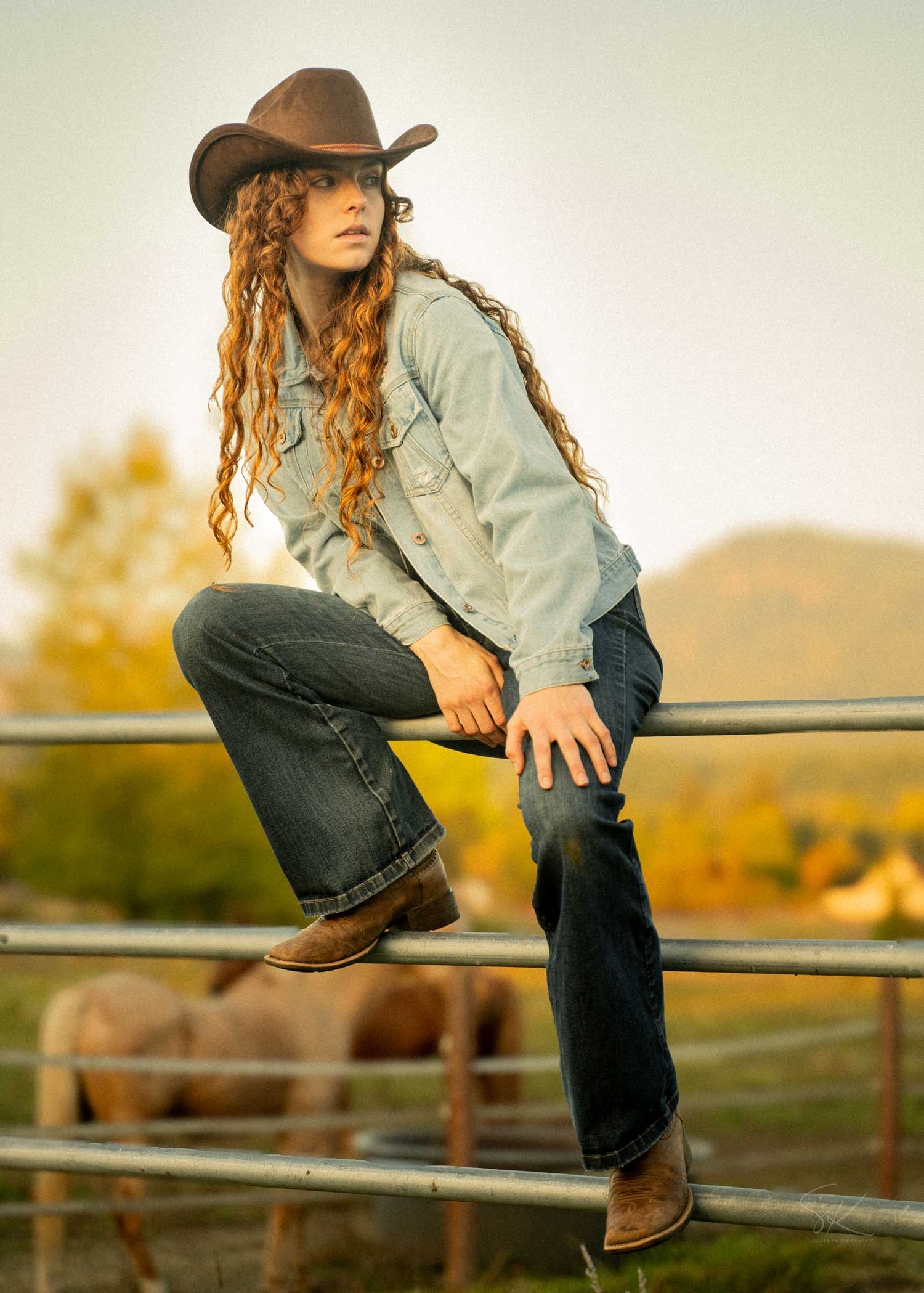 Young woman in cowboy hat and jeans sits on a ranch fence with curly hair and boots, livestock visible behind her.