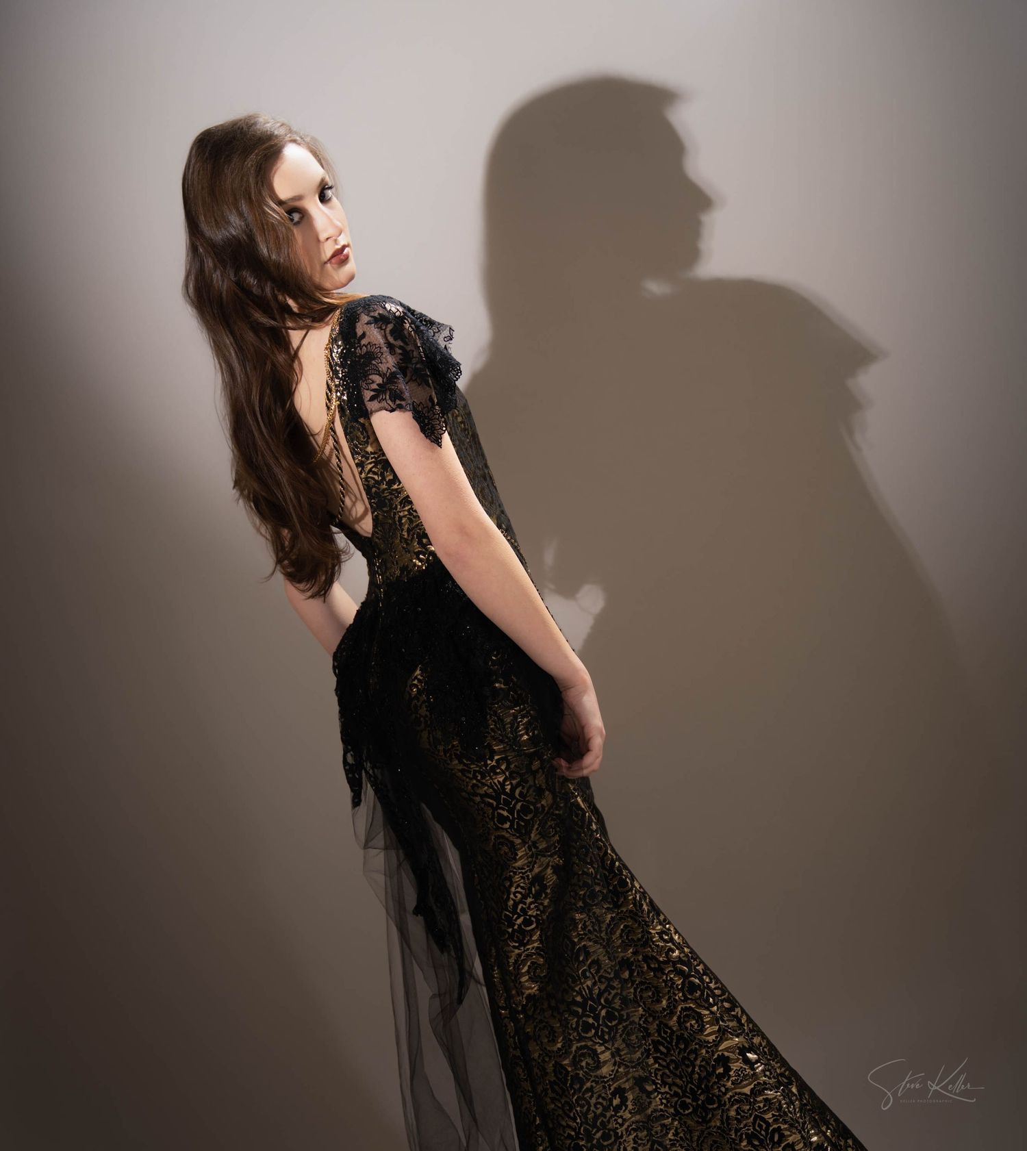 A model poses in an elegant black lace evening gown with dramatic shadow on a neutral backdrop.