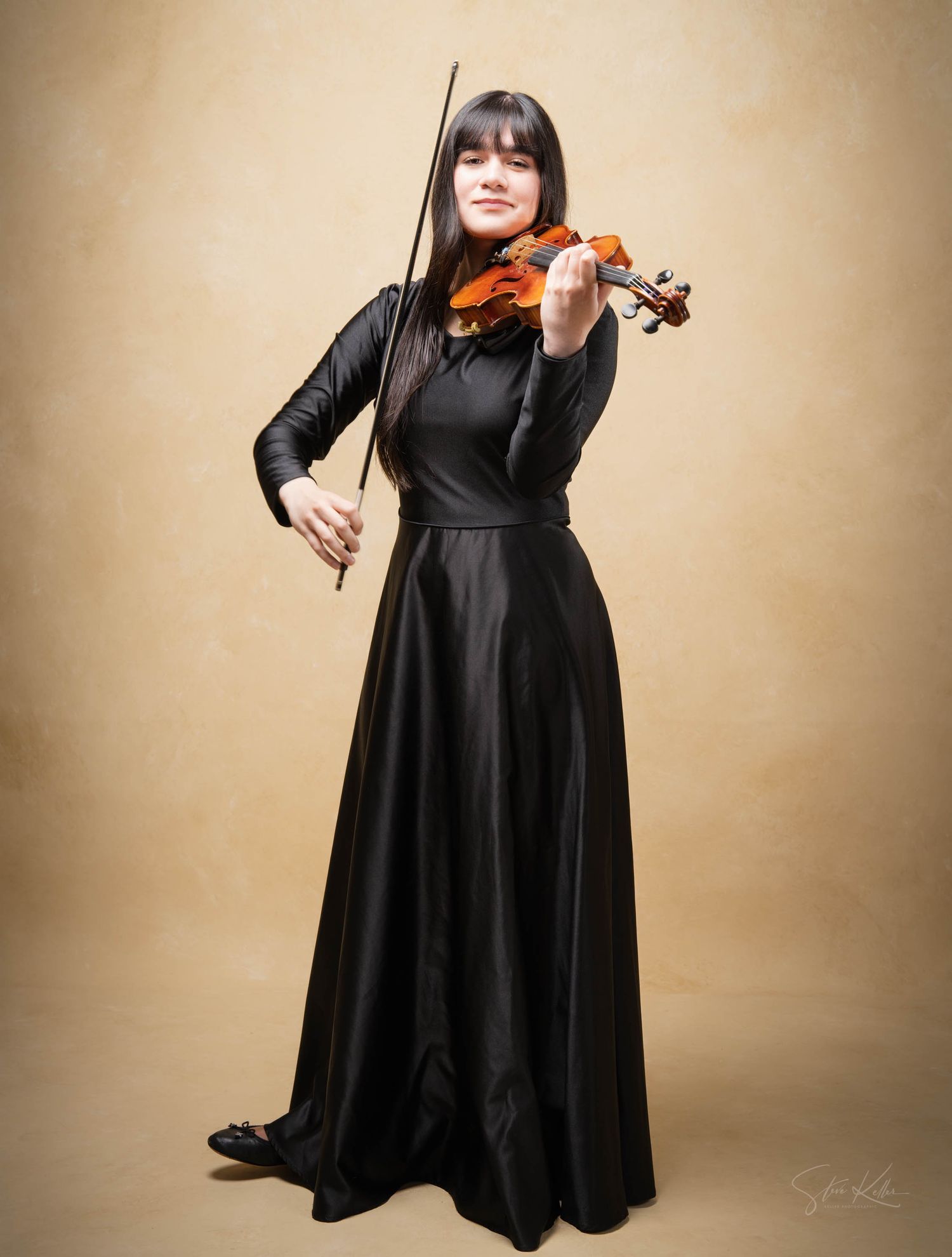 Musician in flowing black dress performs with violin against beige backdrop during elegant portrait session.