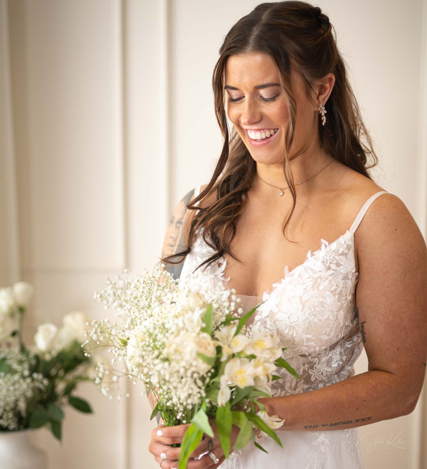 A bride in a white lace gown smiles while holding a delicate bouquet of white flowers and baby's breath.