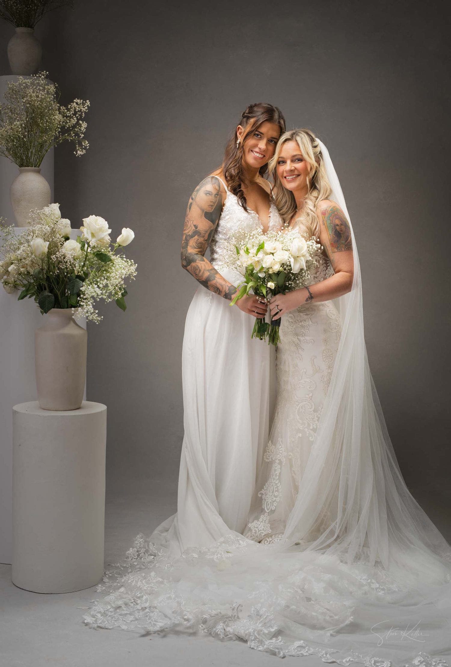 Two brides in elegant white wedding gowns pose together with bouquets against a gray backdrop with floral arrangements.