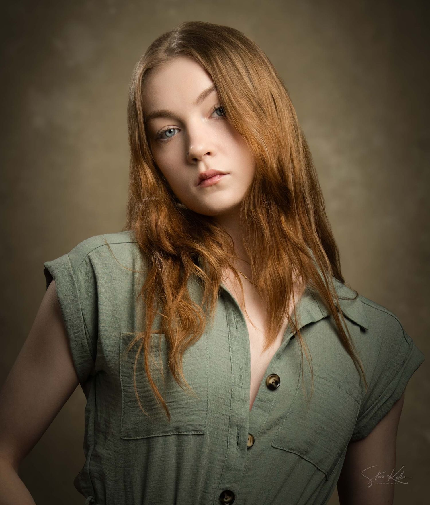 Portrait of a person with flowing red hair wearing a sage green button-up top against a dark moody background.