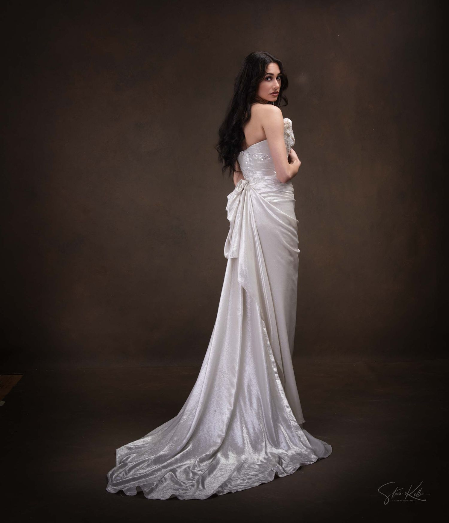 A bride in an elegant white satin wedding gown with a flowing train poses against a dark studio backdrop.