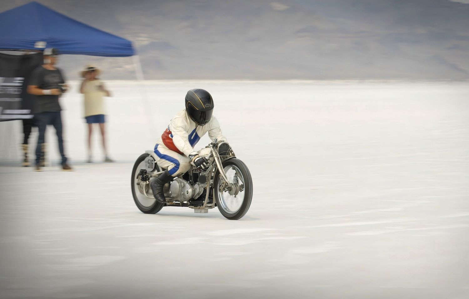 Motorcyclist racing on salt flats at high speed during a land speed record event with spectators in background.