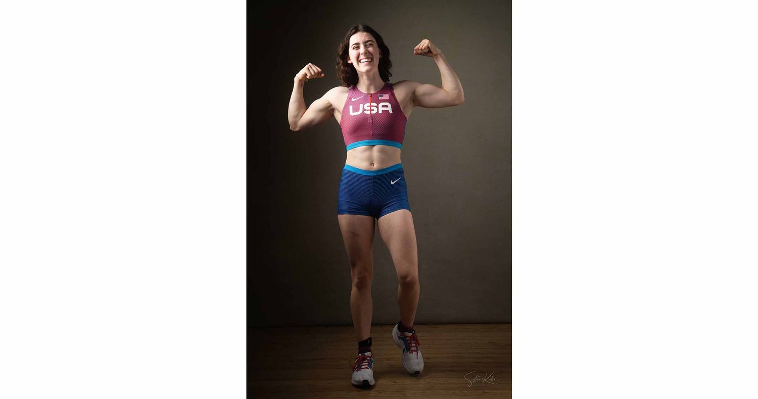 Female track athlete in a USA crop top and blue shorts flexes both biceps while smiling in a studio portrait.