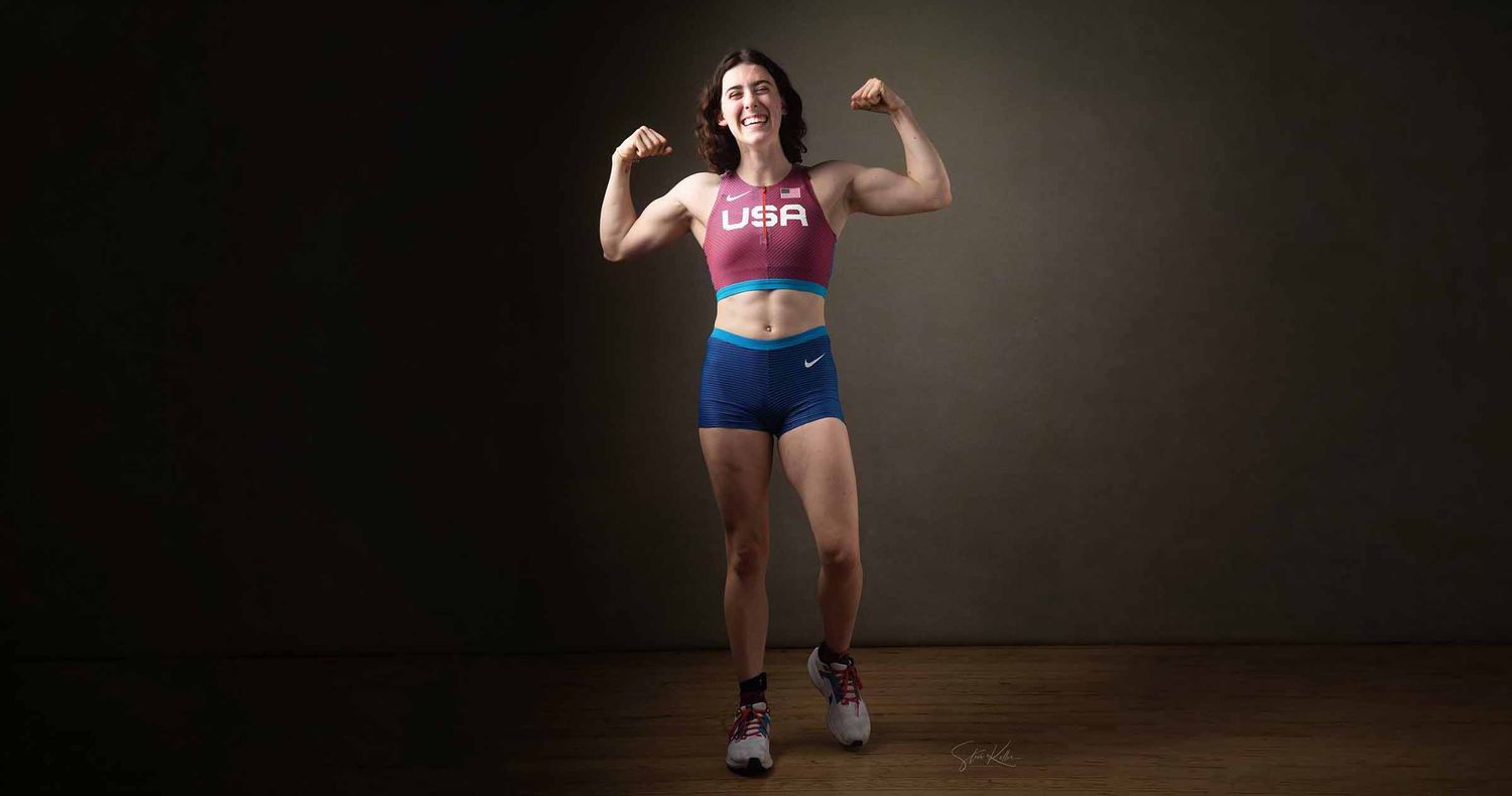 Female athlete in a USA crop top and blue shorts flexes both biceps while smiling against a dark studio backdrop.