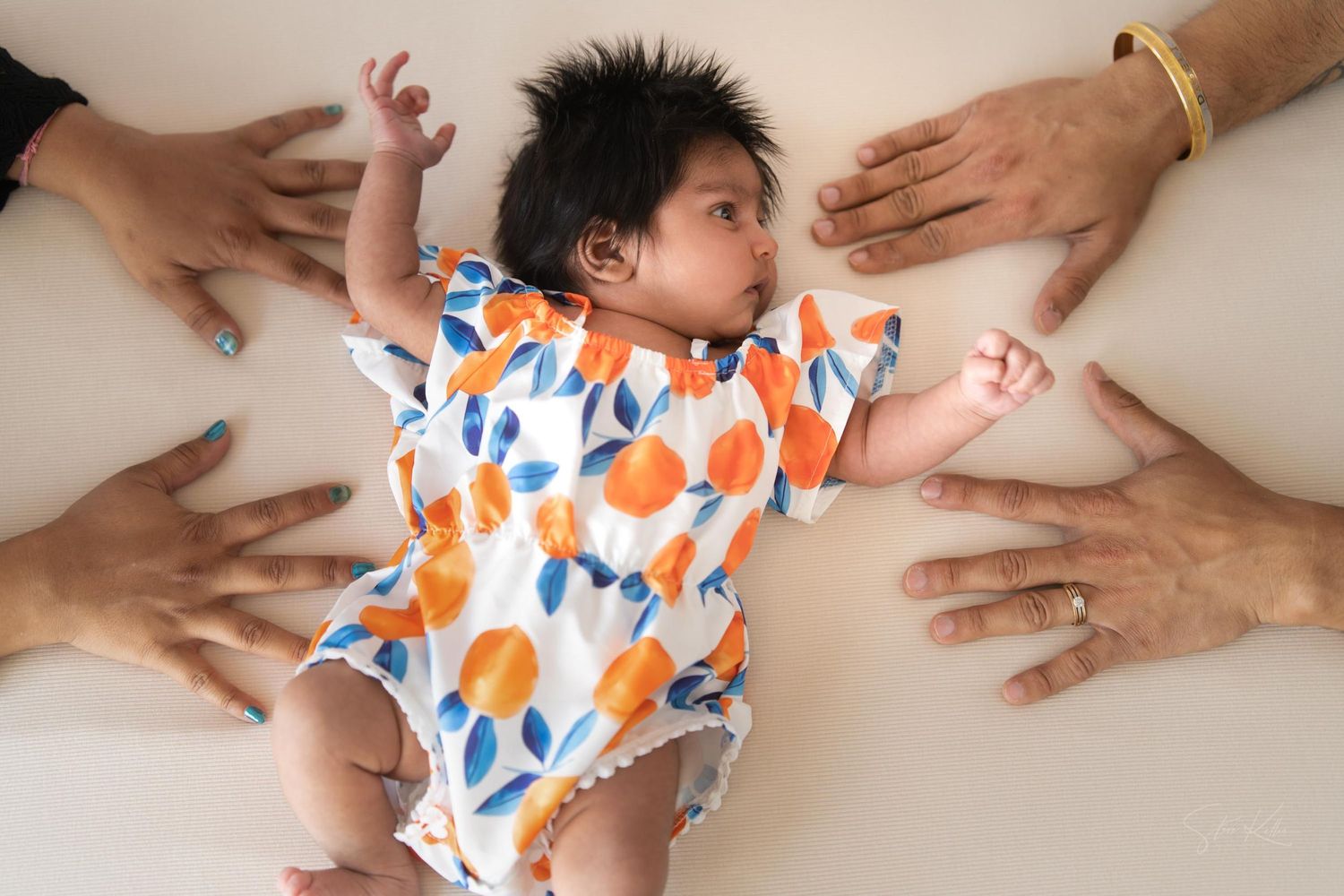 Baby in colorful orange and blue patterned onesie lies surrounded by reaching hands on white background.